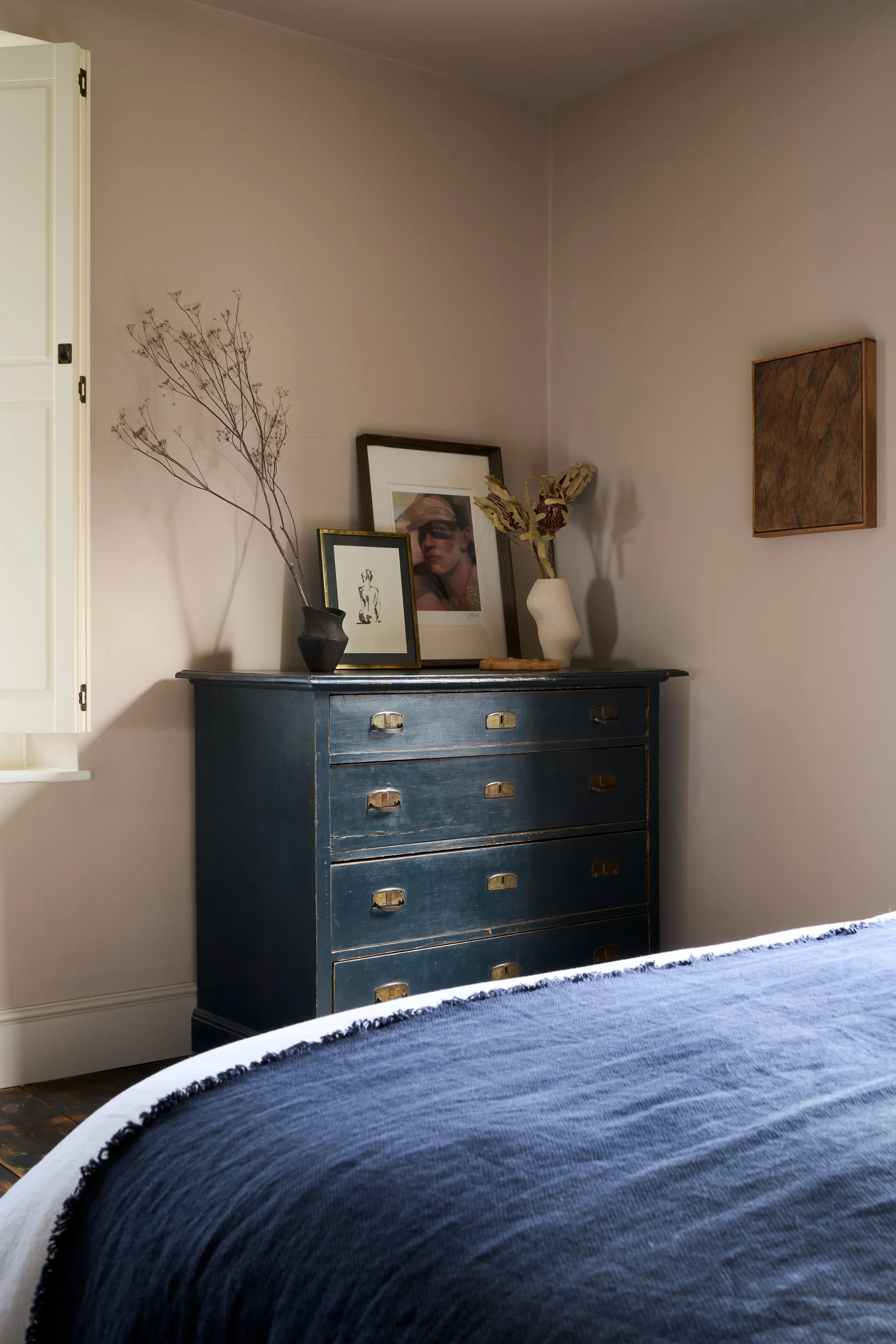 Bedroom showcasing a masterclass in rustic refinement and traditional heritage. The composition balances a distressed antique painted chest of drawers with tactile fringed bed blue linen and artisan ceramics, layered alongside contemporary framed art