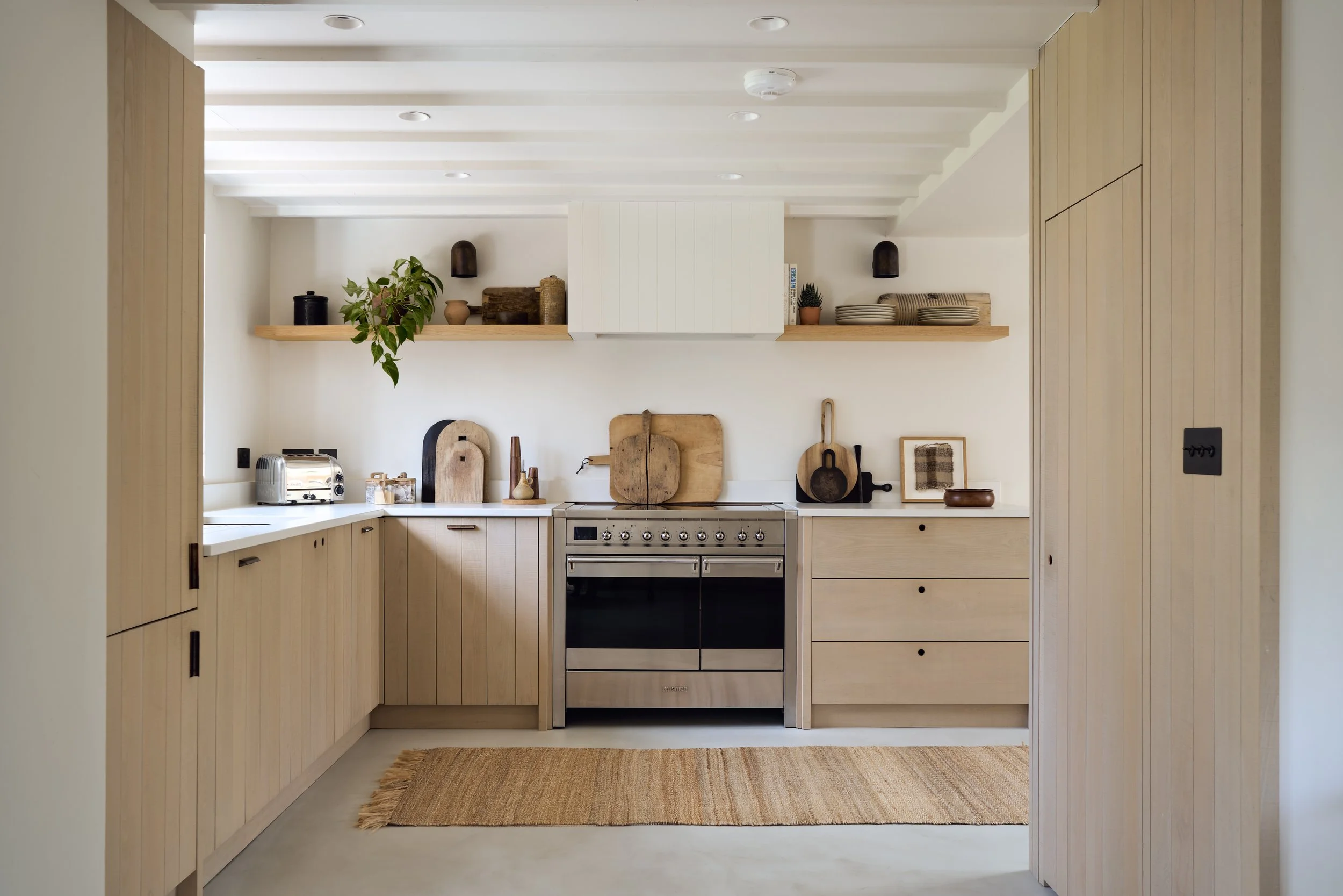 Professional architectural photograph of a bright, minimalist kitchen featuring natural light oak vertical panelled cabinetry, white countertops, and a stainless steel oven.