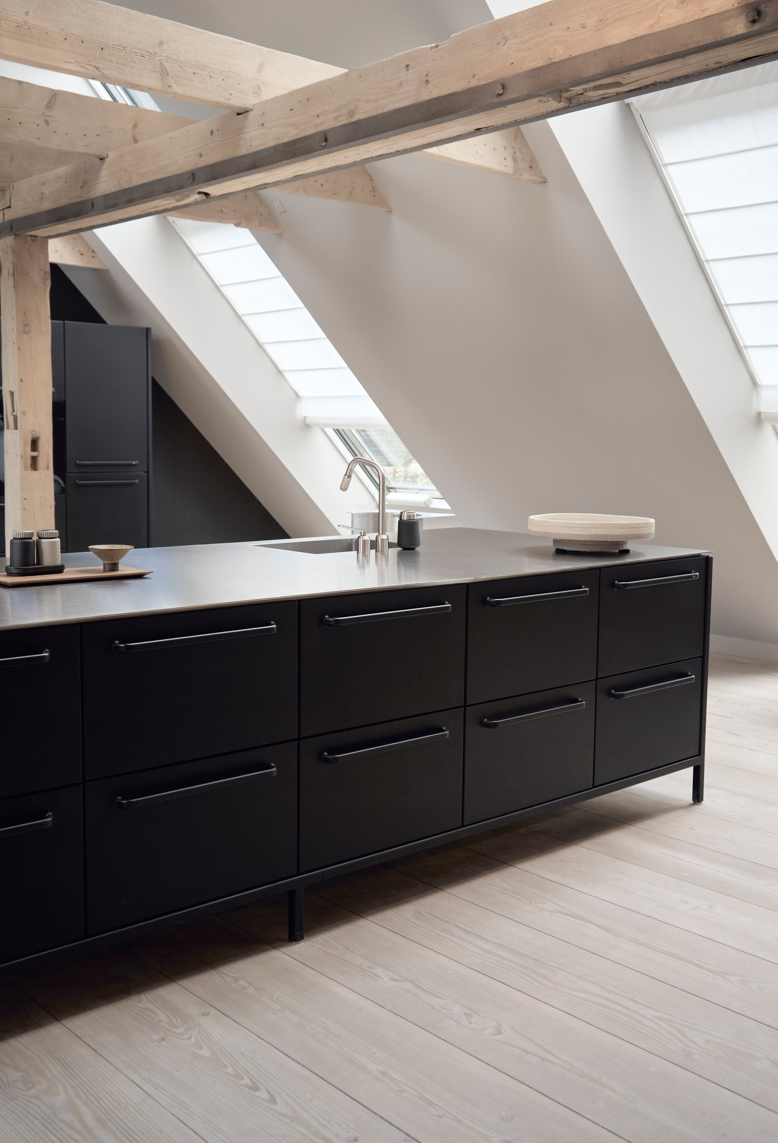 Modern black Vipp kitchen island with stainless steel countertop in an attic loft featuring exposed rustic wooden beams, skylights, and light-toned wooden floors. 