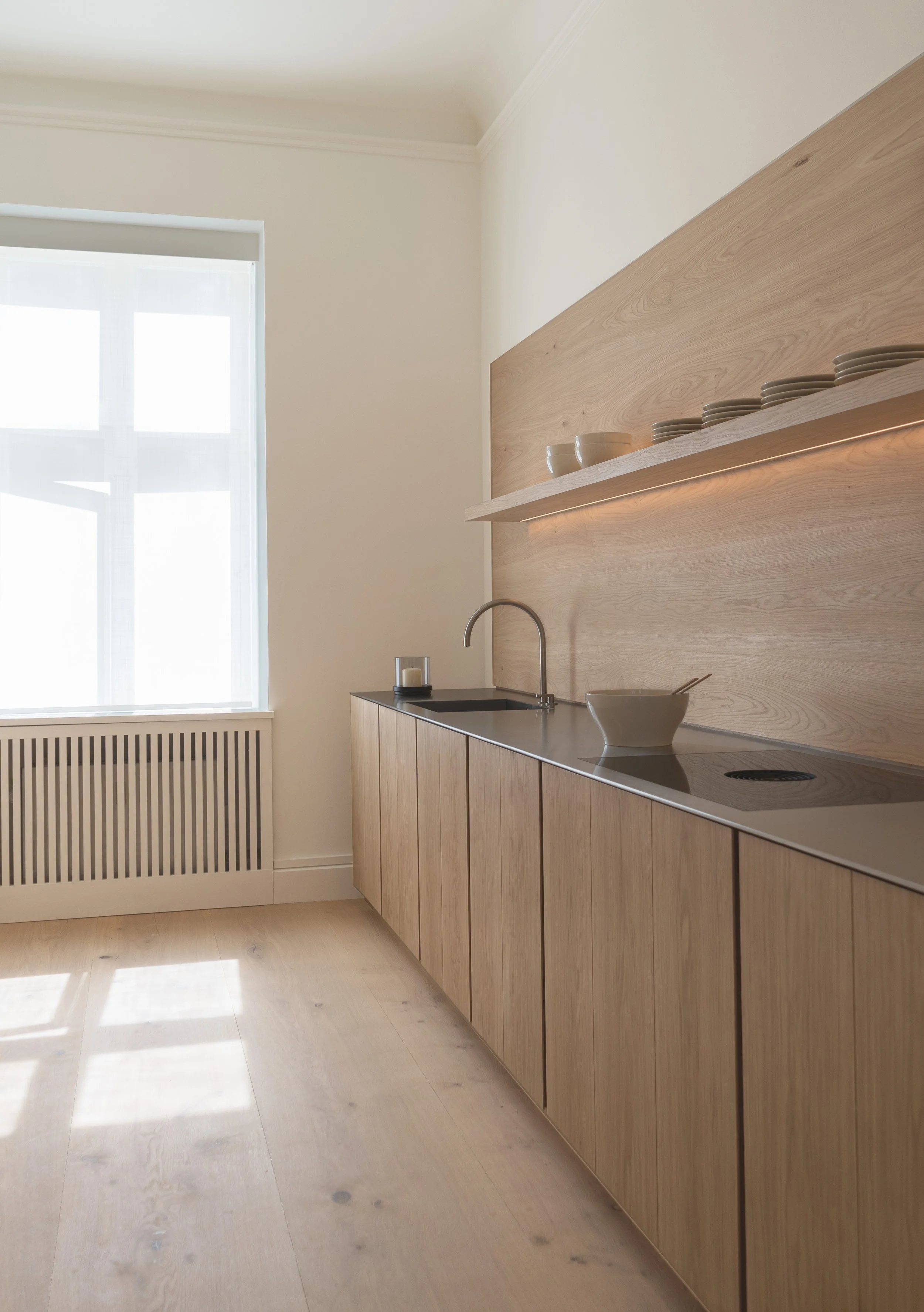 Minimalist kitchen in the Dinesen apartment Copenhagen by John Pawson, featuring light oak cabinetry, a stainless steel countertop, and integrated LED shelf lighting. 