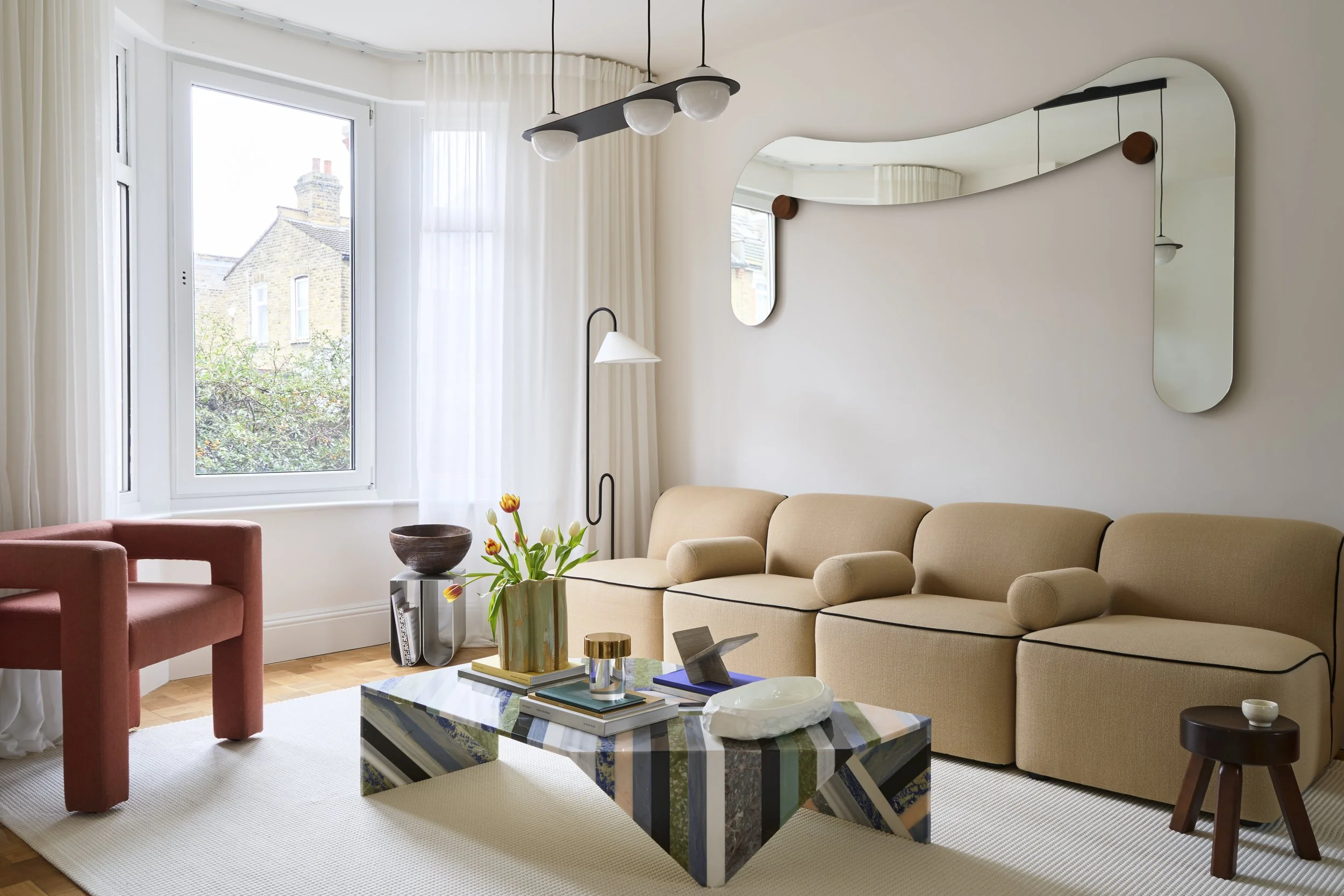 Modern minimalist living room with a beige curved sectional sofa, colourful striped marble coffee table, and an organic wavy mirror. 