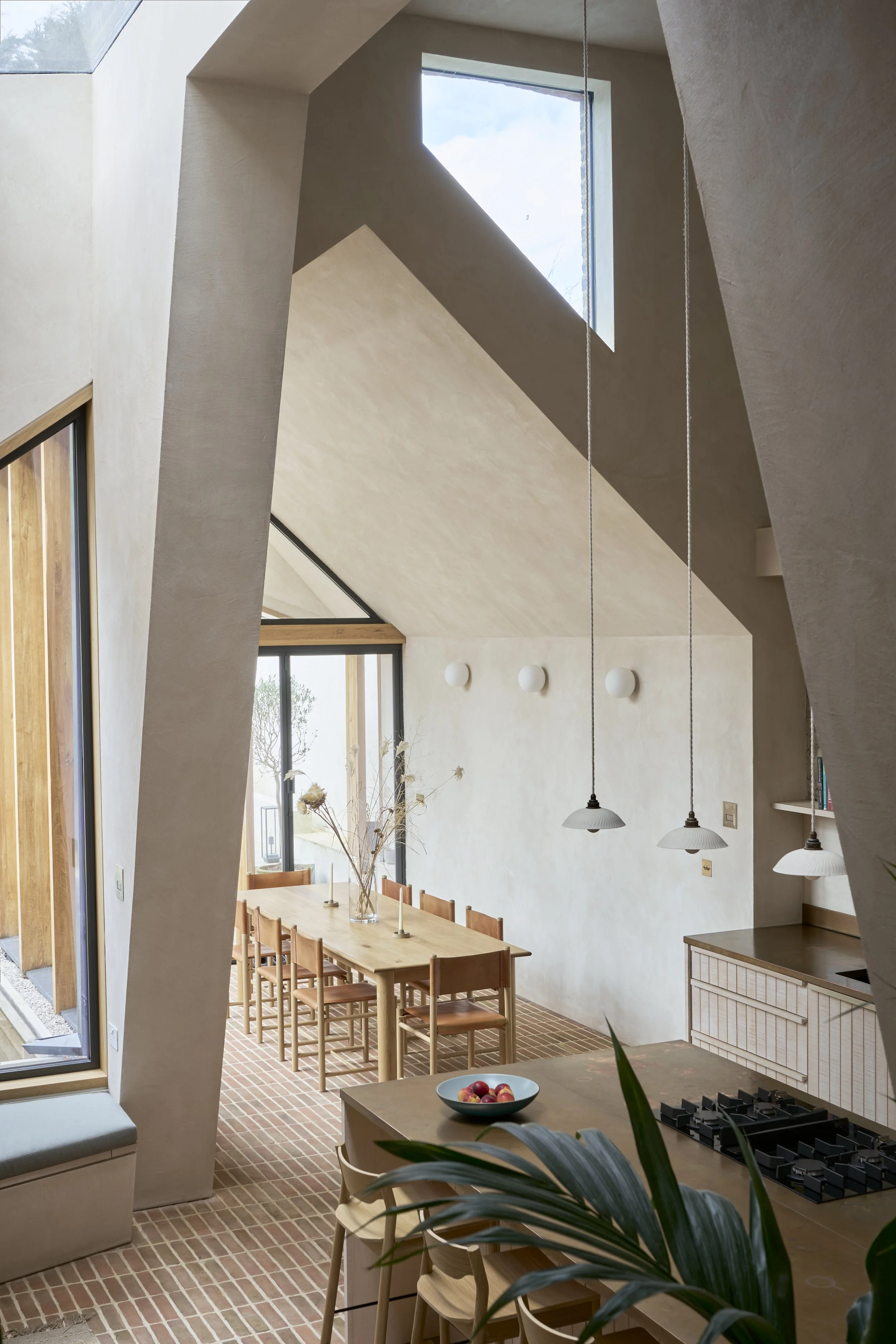 Architectural interior of a modern double-height kitchen-dining room featuring vaulted ceilings, exposed plaster walls, a long wooden dining table, and red brick flooring. 