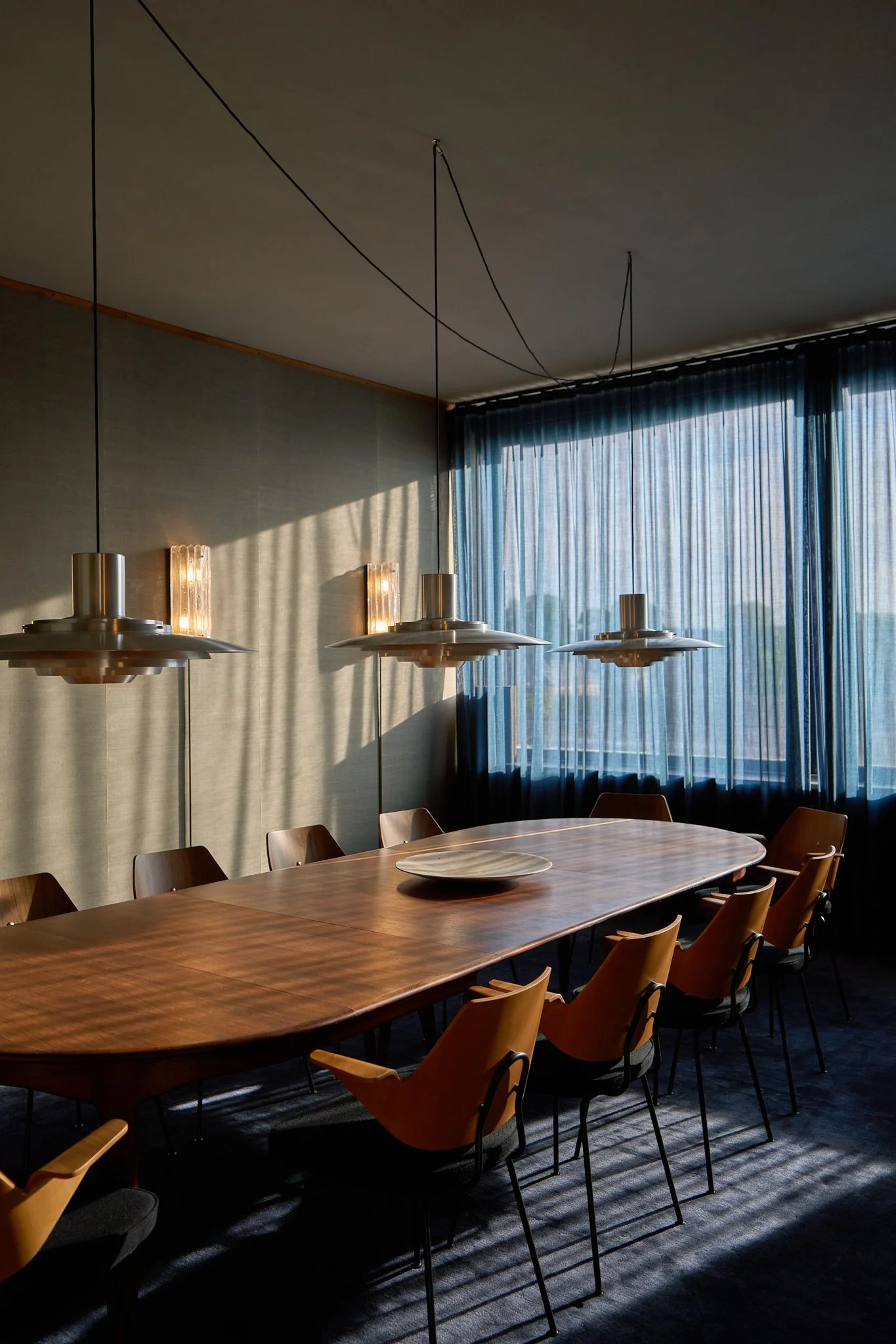 Interior architectural photograph at the Langelinie Pavilion in Copenhagen, showcasing a mid-century boardroom interior by &Tradition. Dramatic, low-angled afternoon sunlight streams through sheer navy blue curtains, casting strong linear shadows acr