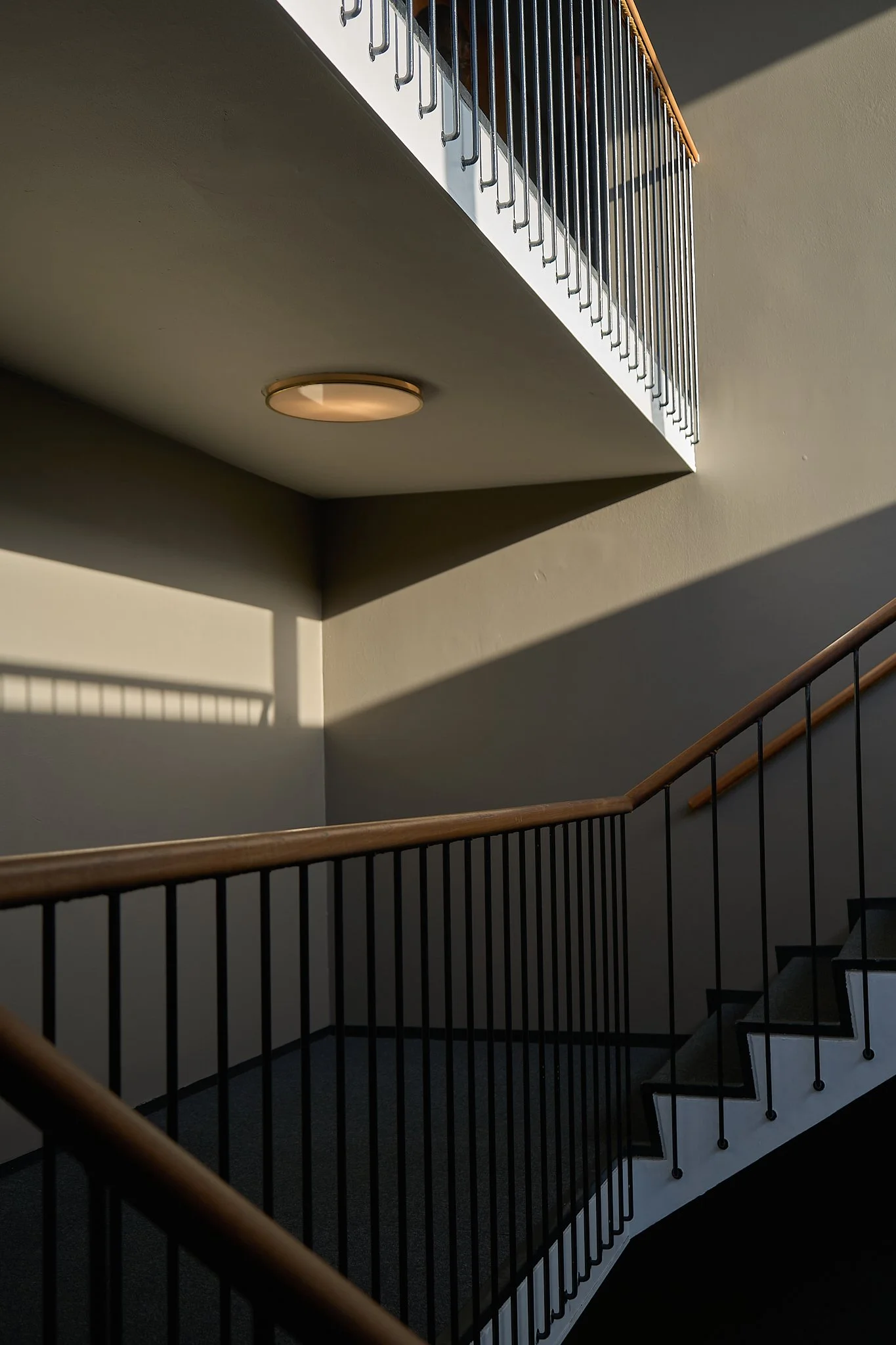 Architectural detail capturing the graphic perspective of an modernist staircase. Crisp, low-angled evening sunlight slices across neutral plaster walls and a round flush ceiling light, creating a high-contrast interplay of linear shadows and minimal