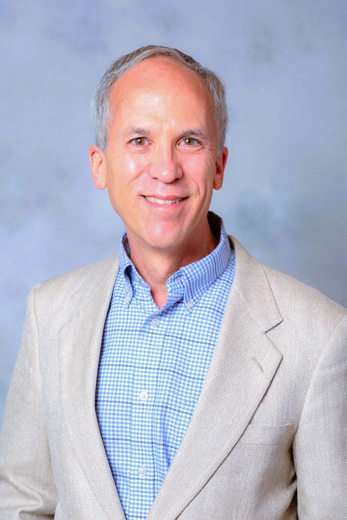 White male with gray hair and brown eyes wearing blue-and-white checked shirt and beige sports jacket with light blue background