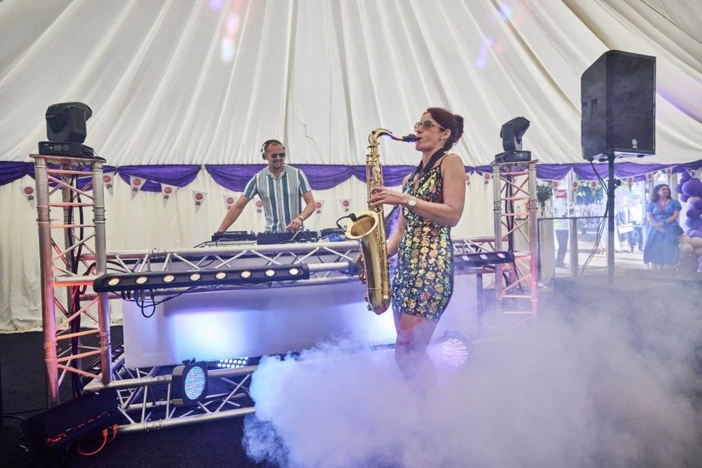 Woman playing saxophone at a DJ event inside a decorated tent with purple and white drapery, fog effects, and a DJ setup in the background.