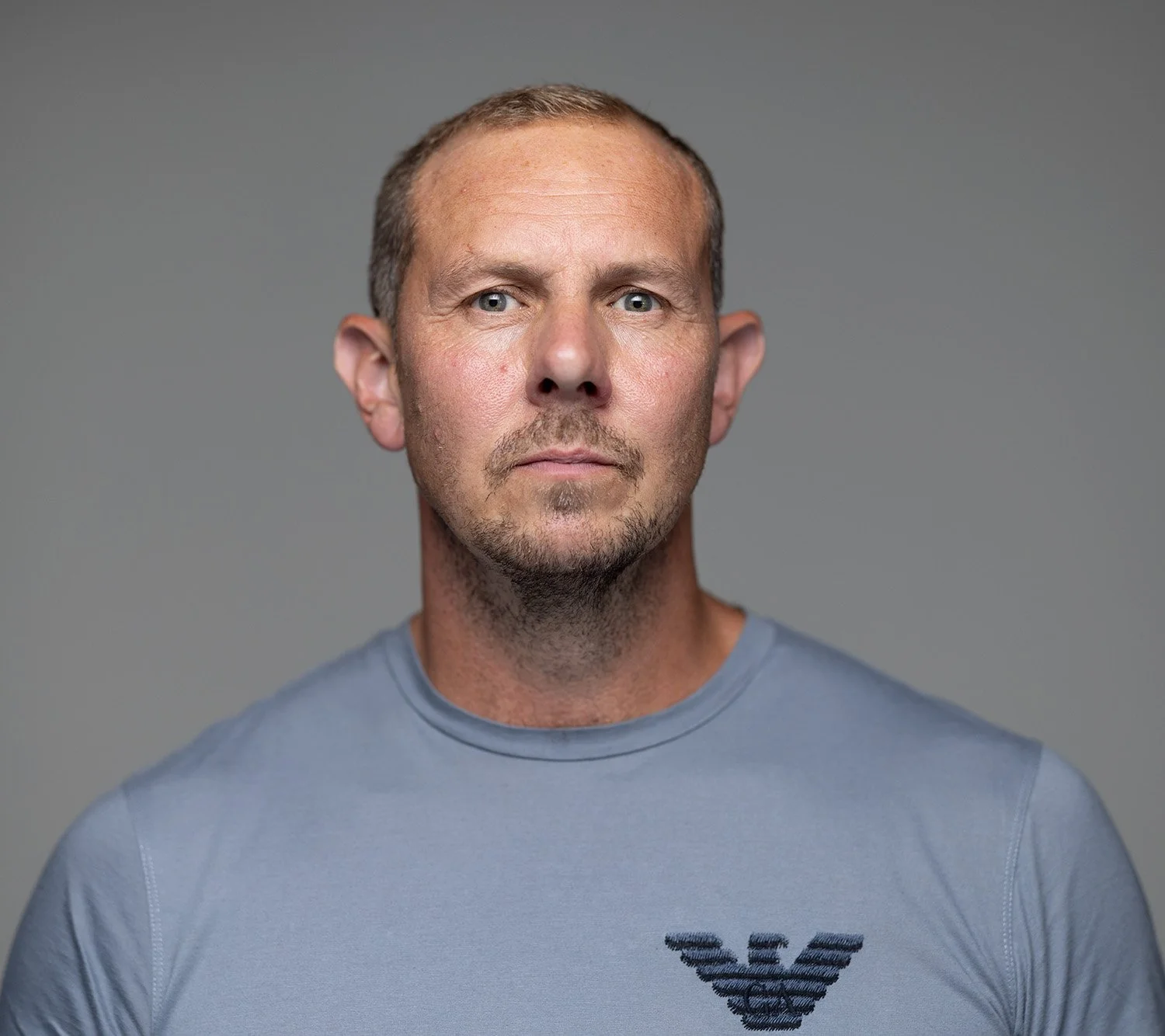 Close-up of a man with short light brown hair, light eyes, and a rugged face, wearing a gray T-shirt with a black logo on the left chest, against a plain gray background.