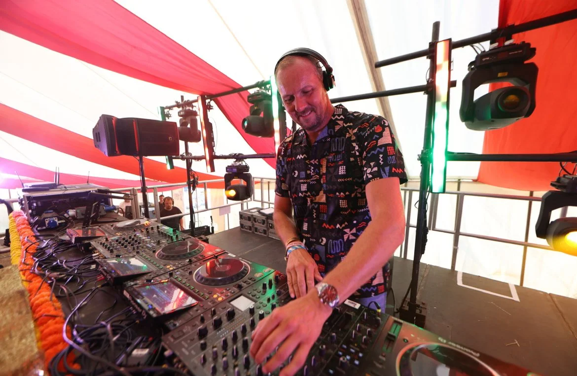 A DJ in a colorful tent, smiling and wearing headphones, mixing music on a DJ console with various equipment and colored lights.