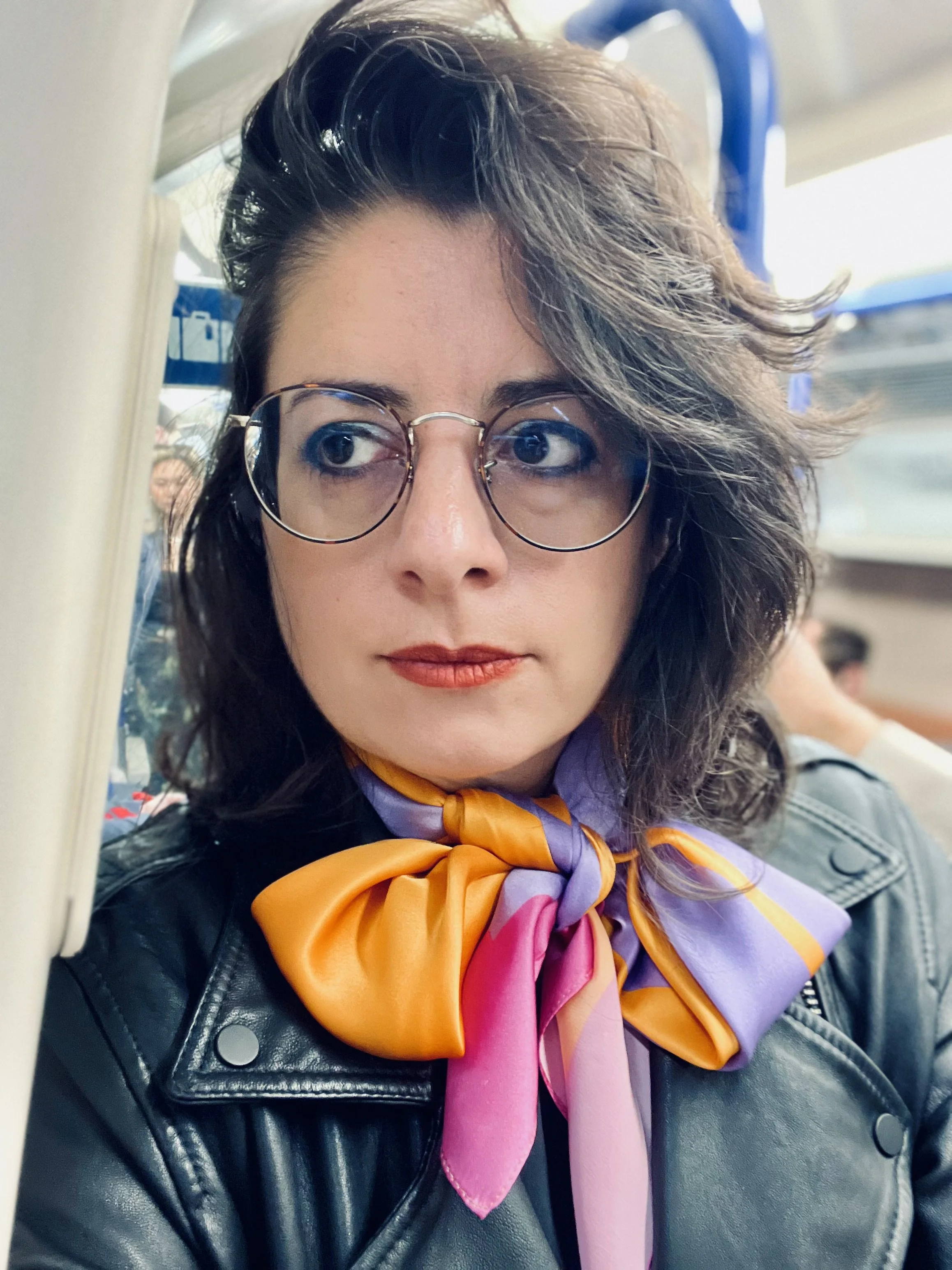 A woman wearing glasses with dark, curly hair styled in waves, dressed in a black leather jacket, with a colorful scarf tied around her neck. She is seated in a public space with a blurred background.