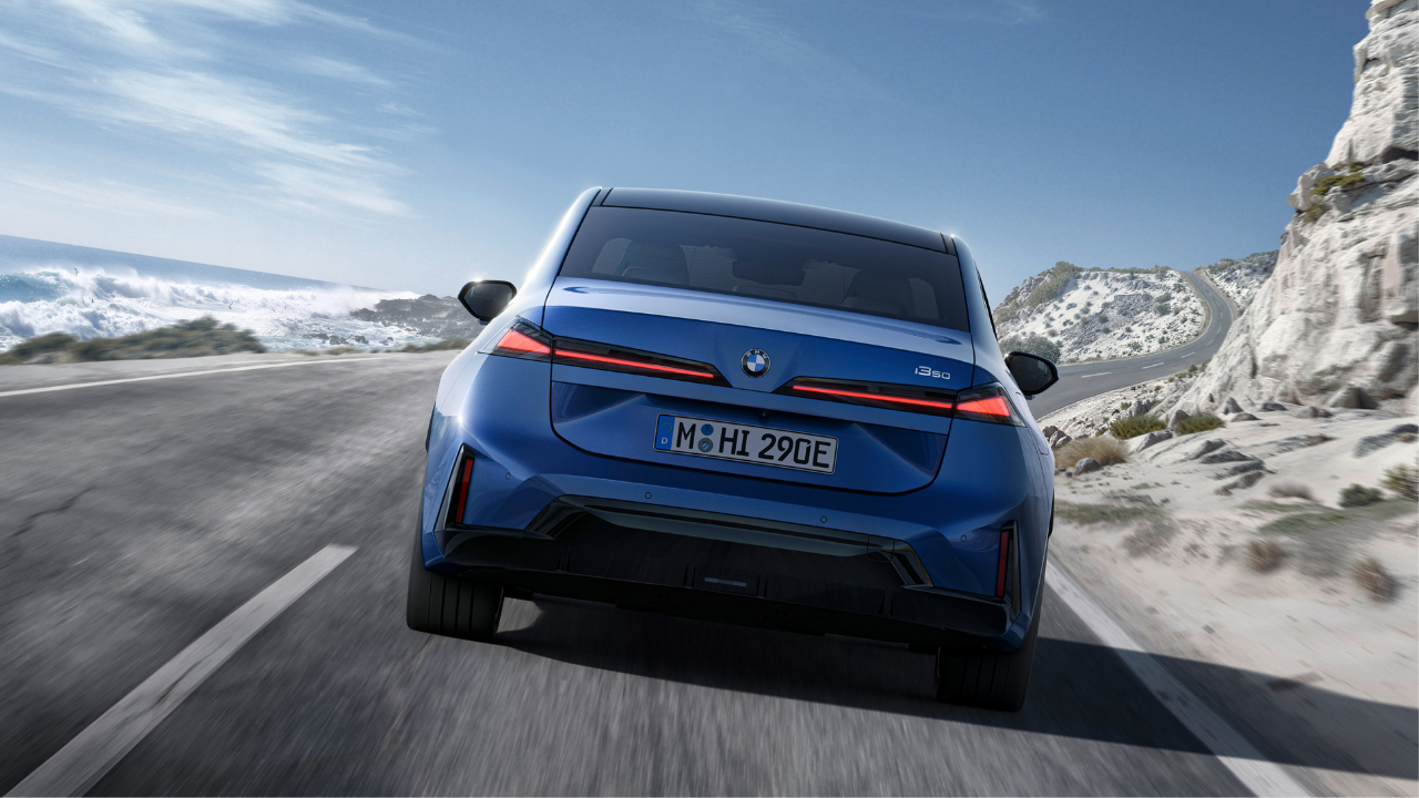 Rendering of a blue BMW i350 driving away on a coastal road, showing sleek EV taillights and sea views.