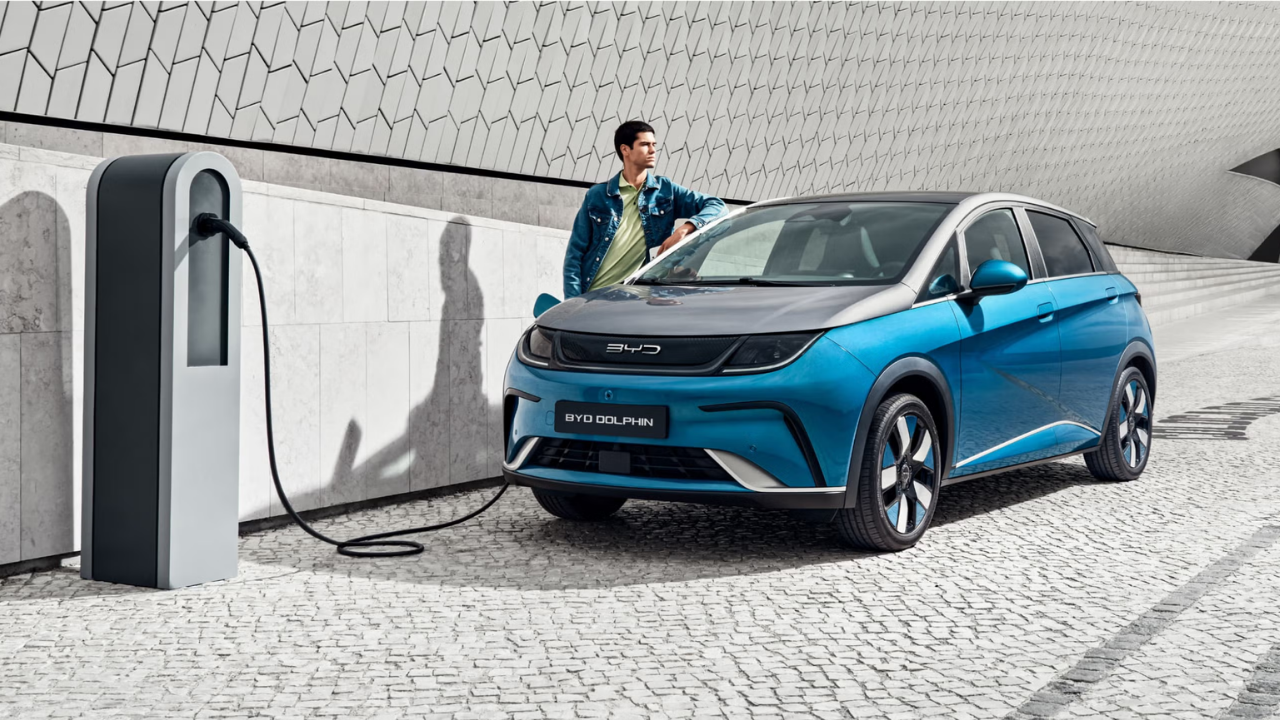 Rendering of a blue and grey BYD Dolphin charging at a modern pedestal station with a man leaning on the hood.