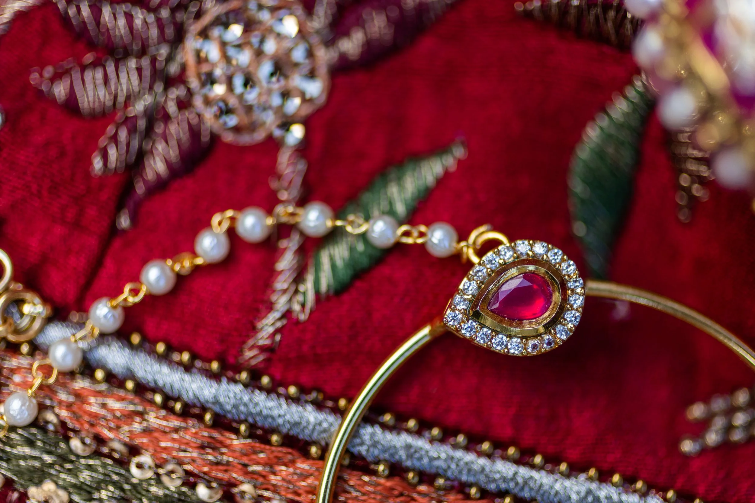 Close-up of jewelry on red fabric, featuring a gold bracelet with a red gemstone surrounded by diamonds, pearl necklaces, and other gold and diamond jewelry.