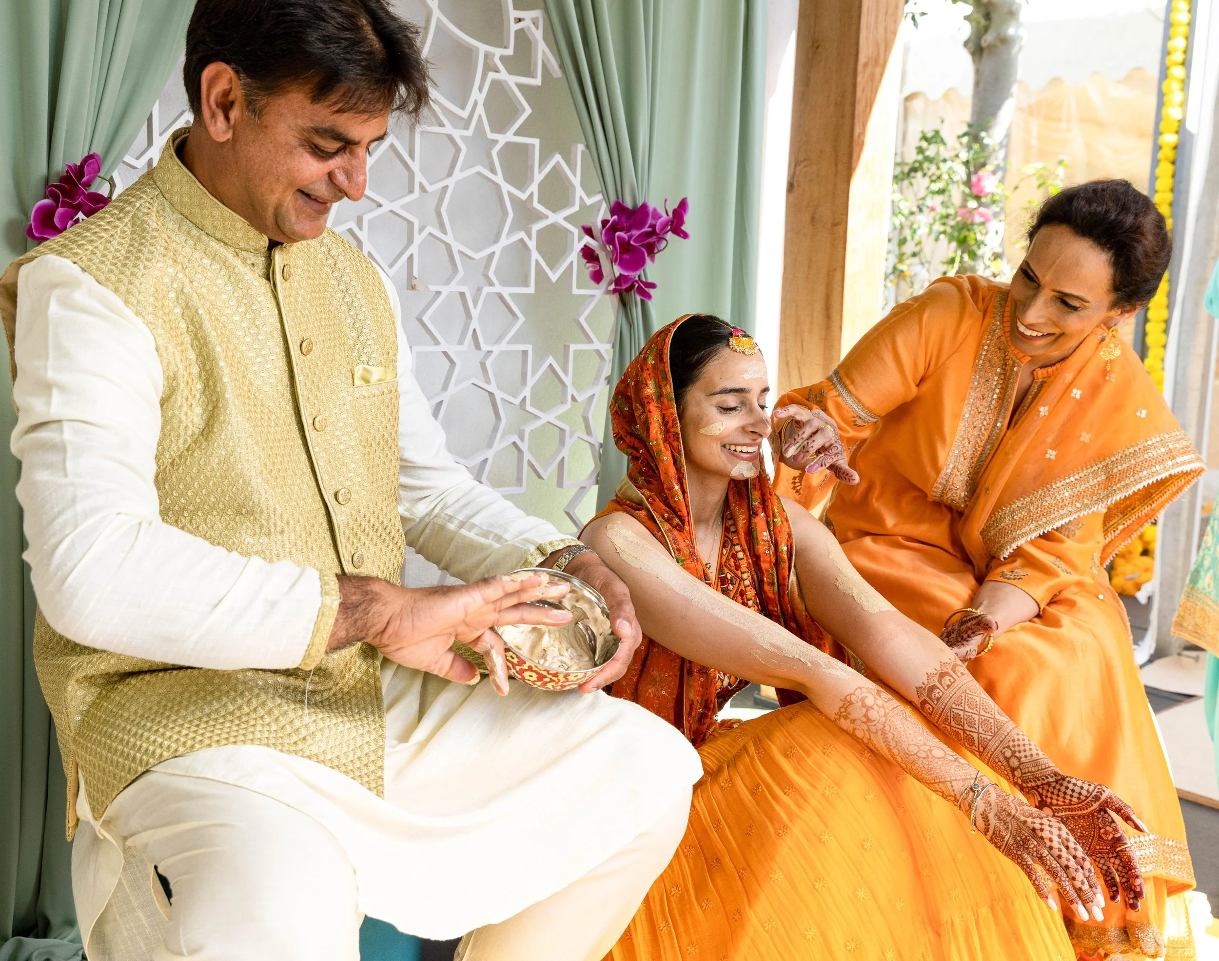 Indian bride smiling as she gets mehndi applied on her arms during her wedding ceremony, with family members helping and celebrating.