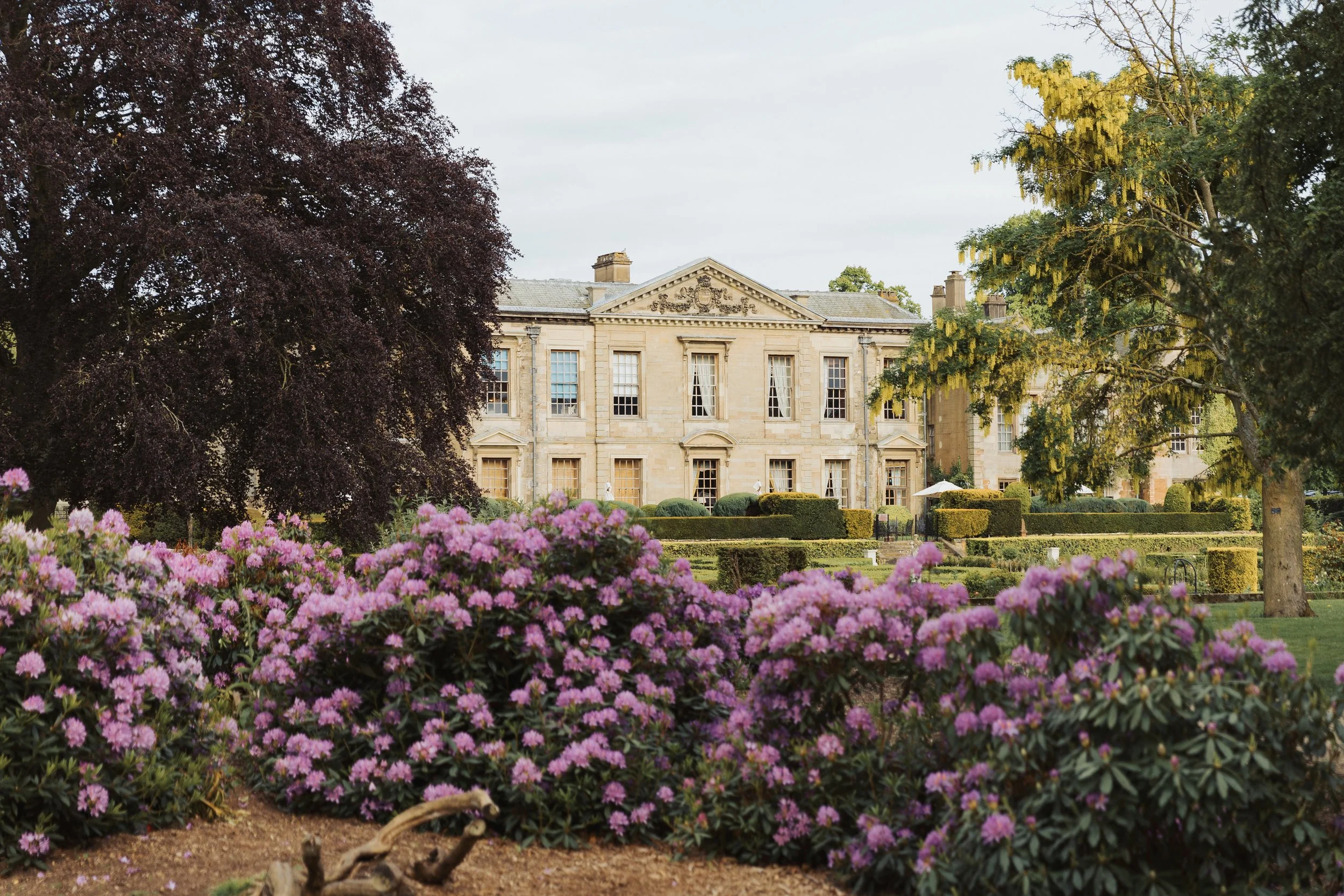 A large, historic mansion with classical architecture, surrounded by a well-maintained garden with pink flowering bushes, trimmed hedges, and tall trees.