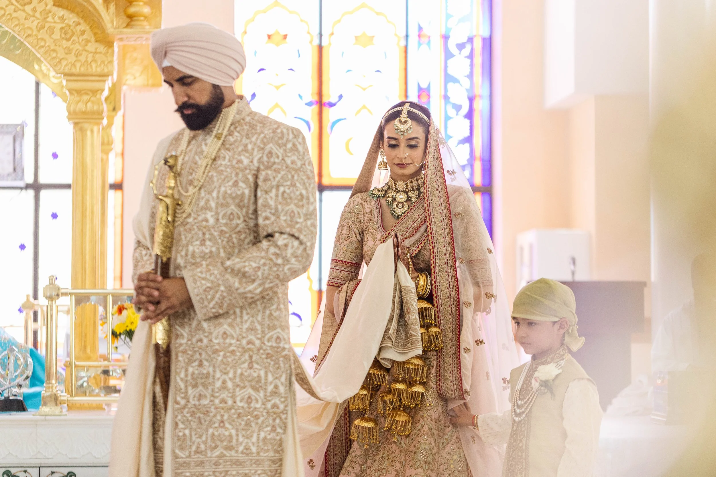 A bride and groom dressed in traditional Indian wedding attire inside a decorated temple, with a young boy also in traditional clothing, standing beside them.