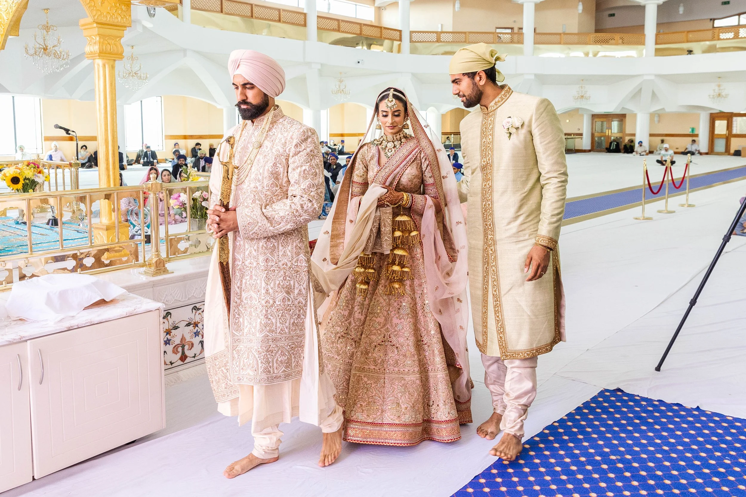 A traditional Indian wedding ceremony with a bride in a pink and gold saree, flanked by two men in traditional attire, inside a spacious hall with guests seated in the background.