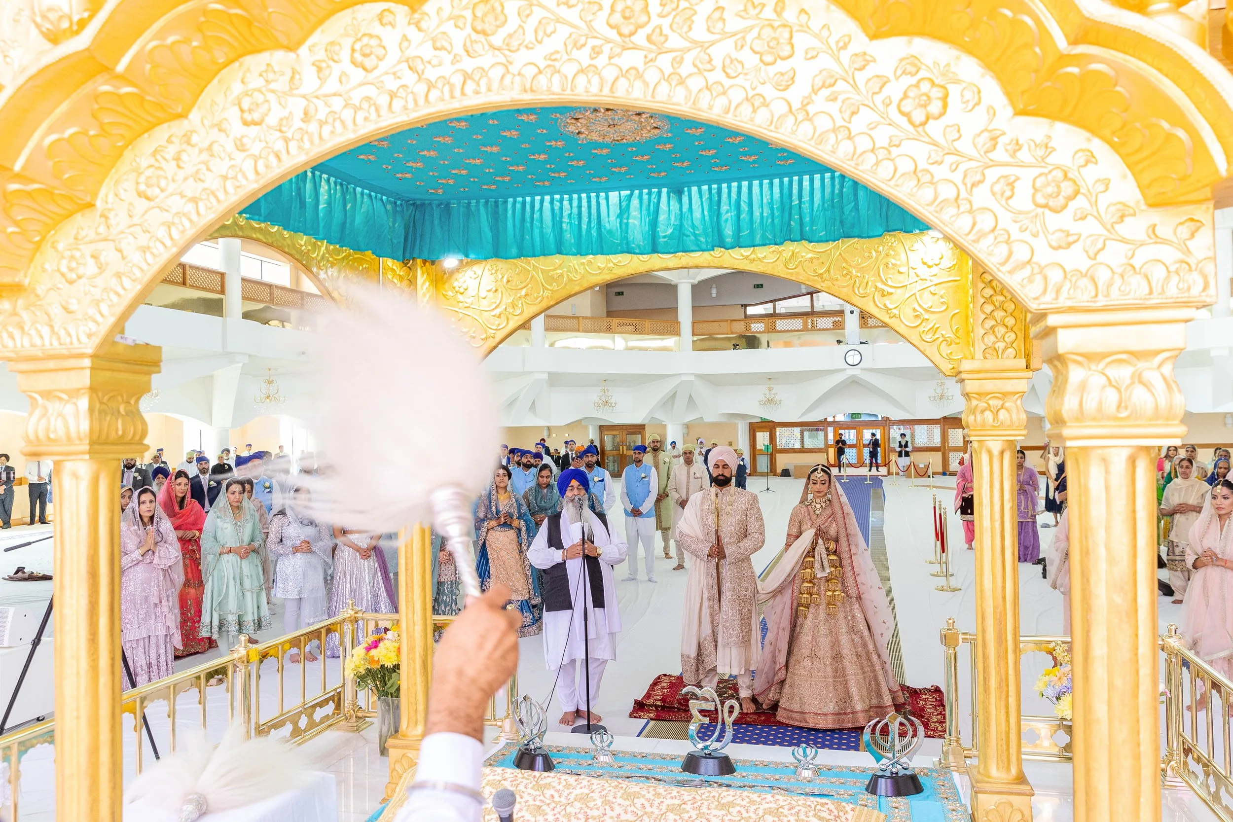 Indian wedding ceremony with bride and groom standing on a decorated platform, surrounded by guests dressed in colorful traditional attire, in a grand hall with gold and turquoise decorations.
