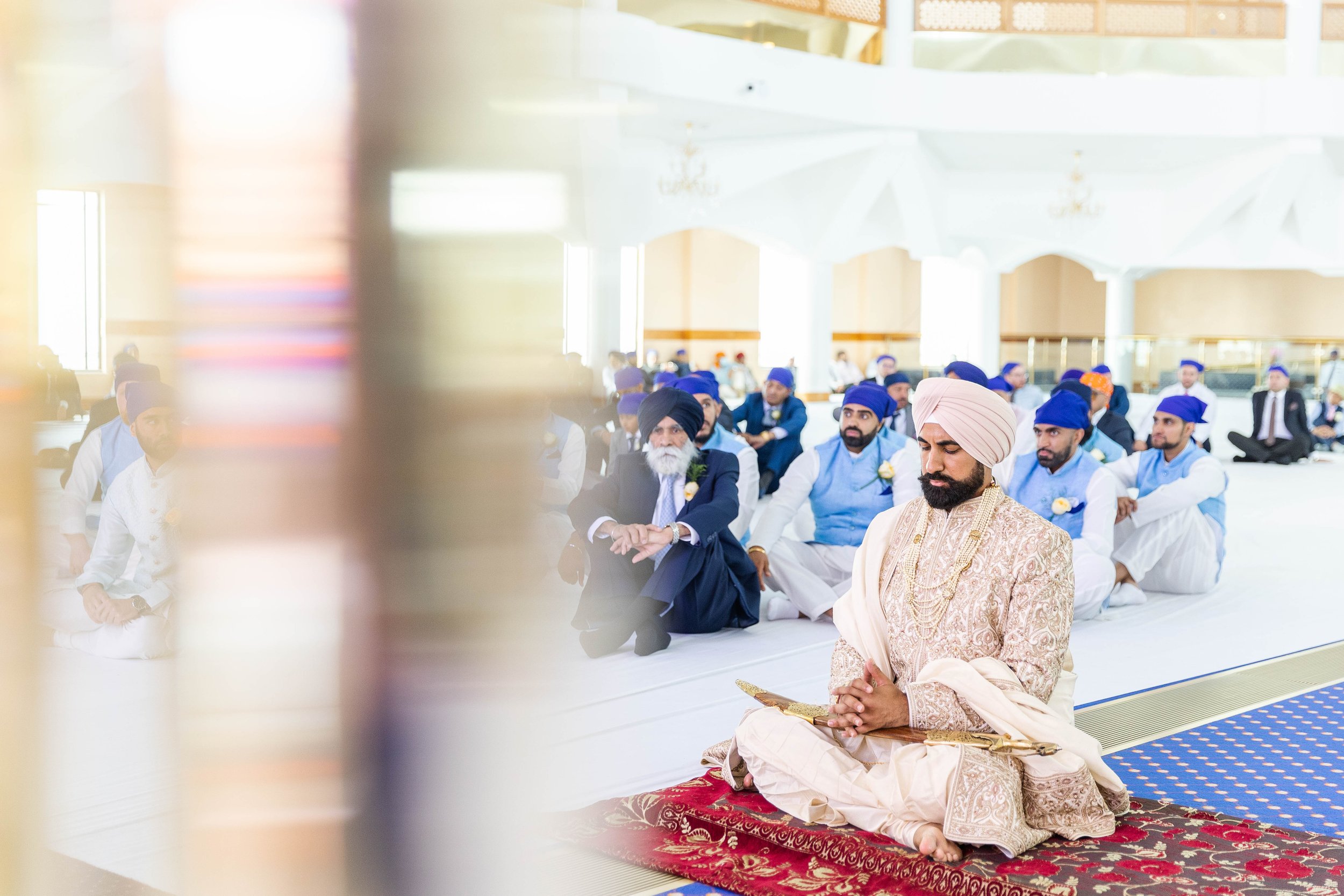 A man in traditional Indian wedding attire, including a cream-colored sherwani and turban, sitting cross-legged with hands clasped in prayer on a decorated red and gold prayer rug. Behind him, a group of men in formal attire including blue vests, whi