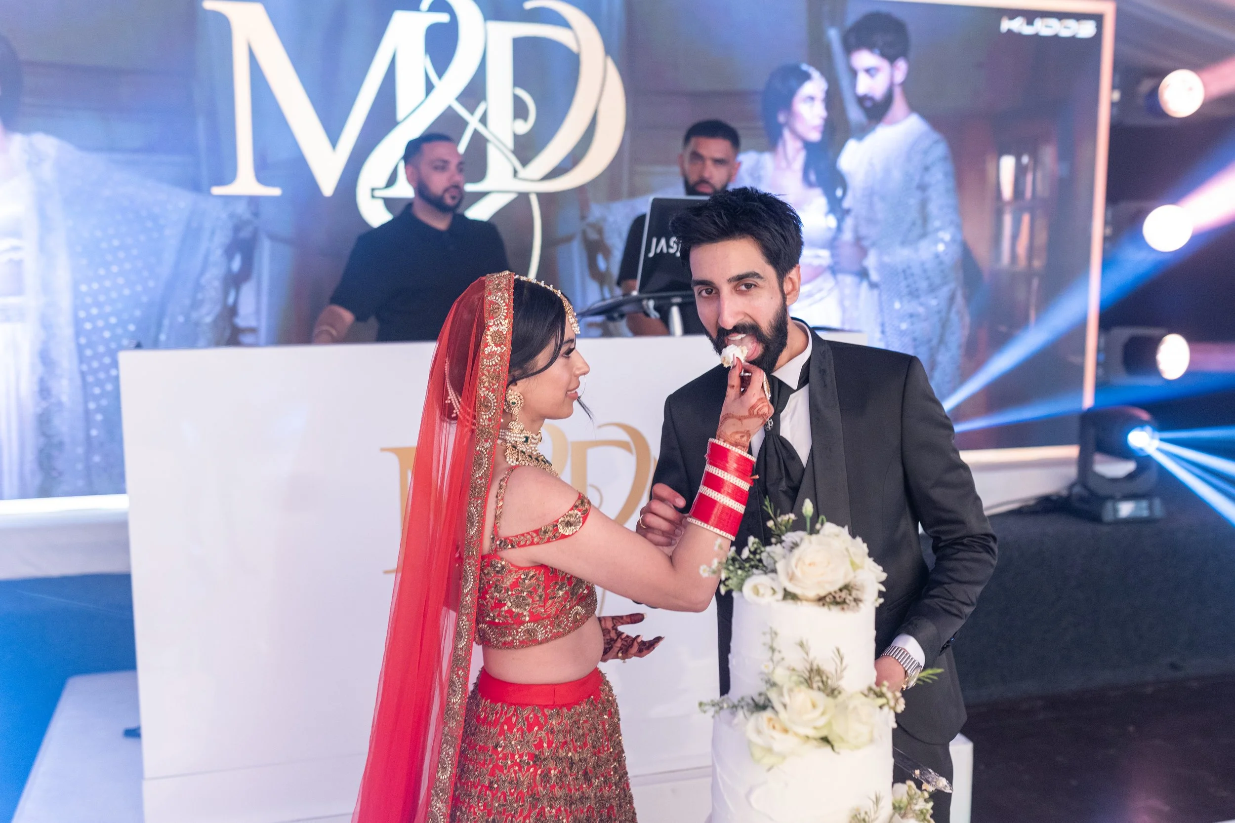A bride in a red and gold traditional outfit feeds cake to a groom in a black tuxedo at a wedding celebration. The groom holds a bouquet of white flowers. In the background, a DJ and a large screen display a couple's photo.