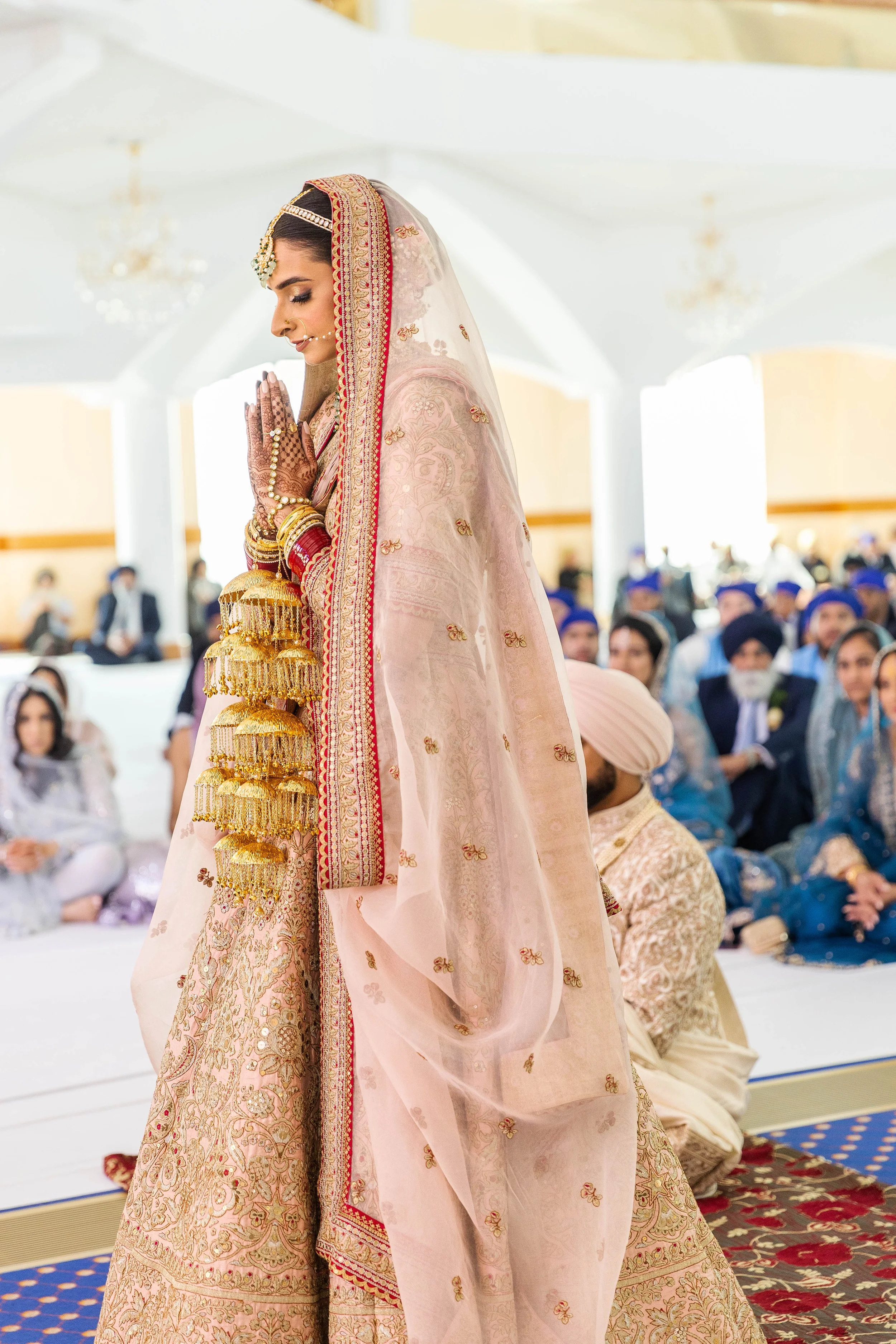 A bride in a traditional pink and gold wedding dress, adorned with jewelry, standing with her hands folded in prayer during a ceremony, surrounded by seated guests in colorful attire.
