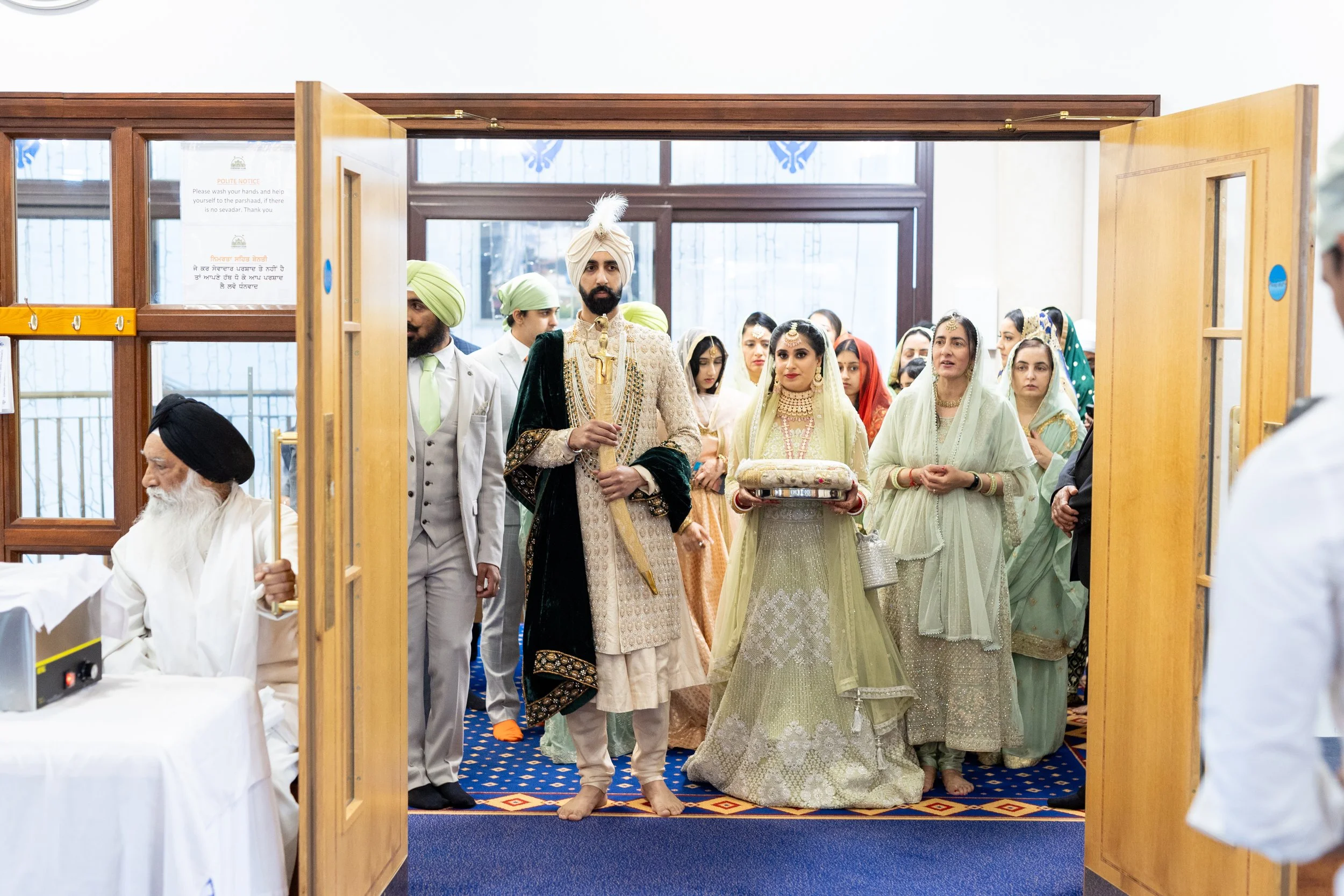 Indian groom and bride in traditional attire walking into a wedding ceremony surrounded by guests, with an elderly man seated on the left and a man on the right in a white shirt.