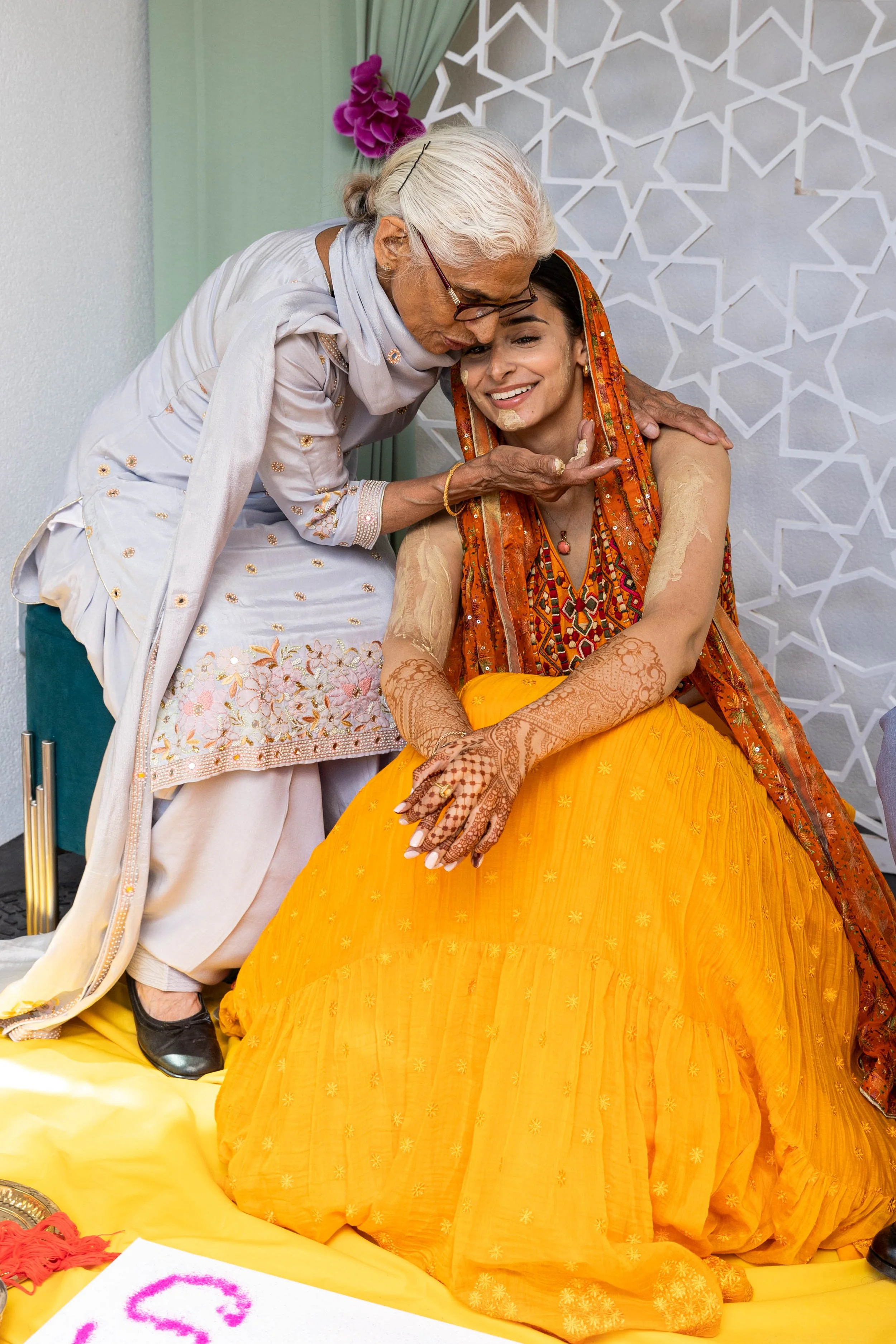 An elderly woman applies a yellow and beige paste to a smiling young woman dressed in orange and red traditional Indian attire, during a cultural or religious ceremony.