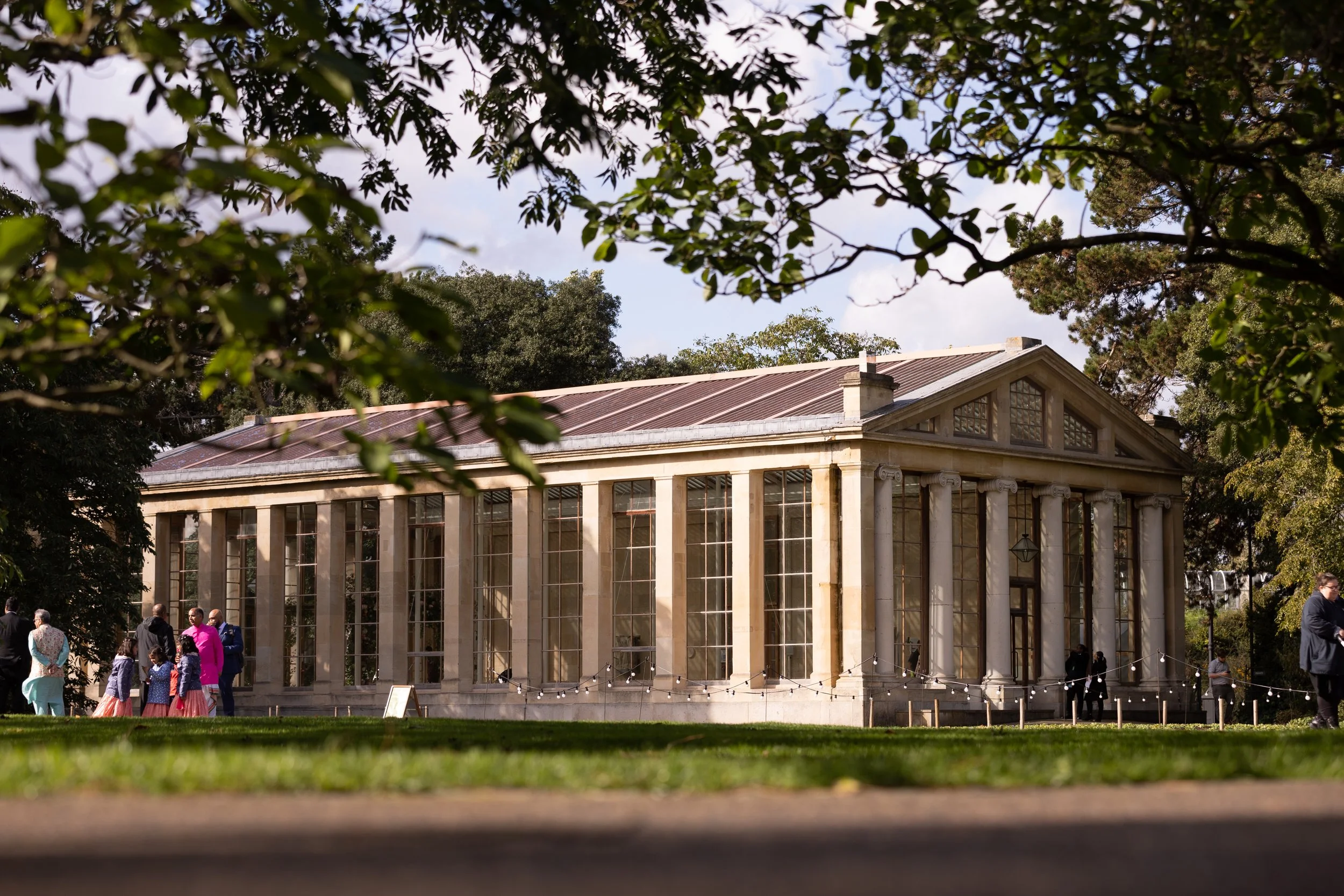 A historic building with tall windows and columns, surrounded by trees, with people standing in front and a string of lights.