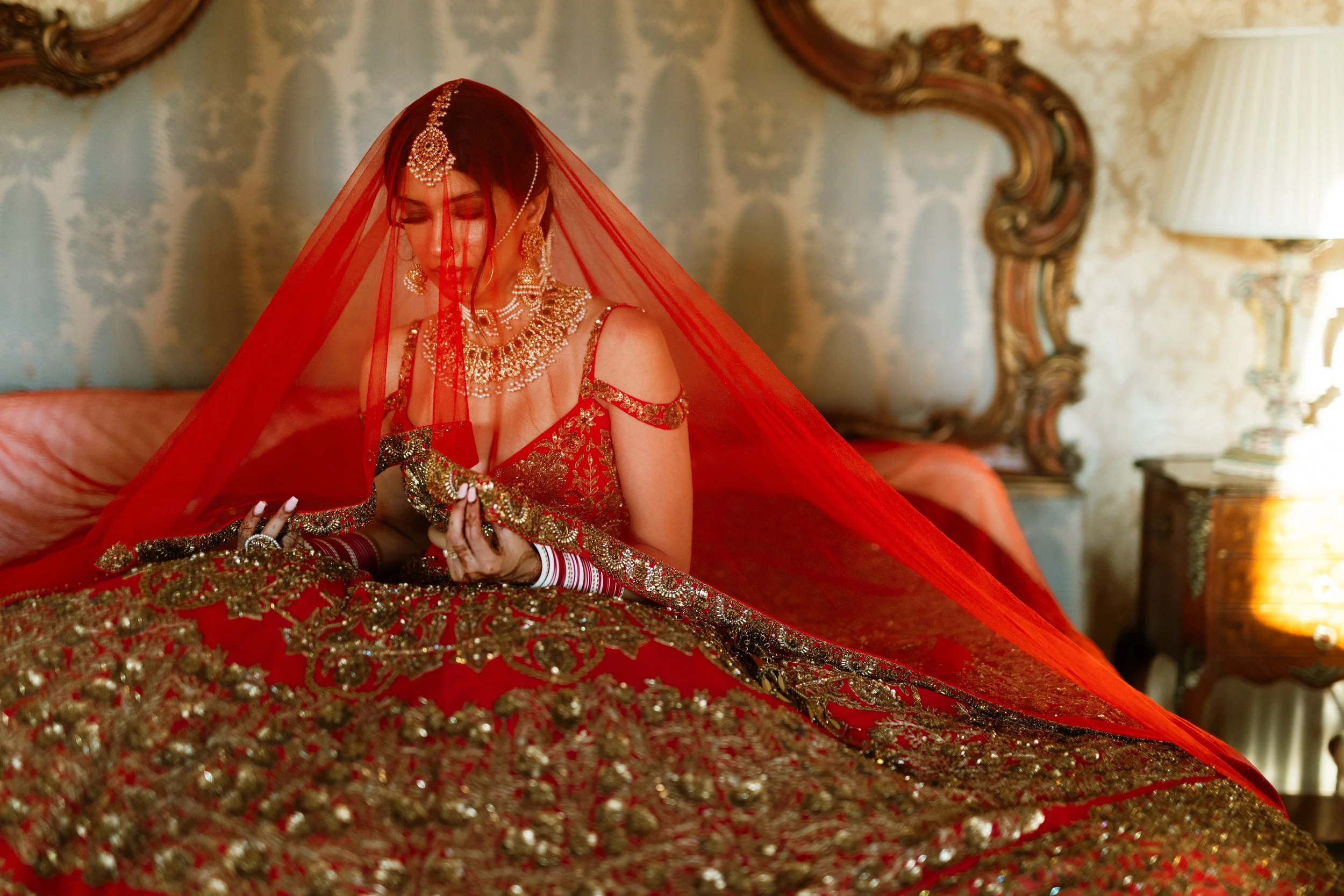 A woman dressed as a bride wearing a red and gold traditional wedding dress, jewelry, and a red veil sitting on a bed in a luxurious room.