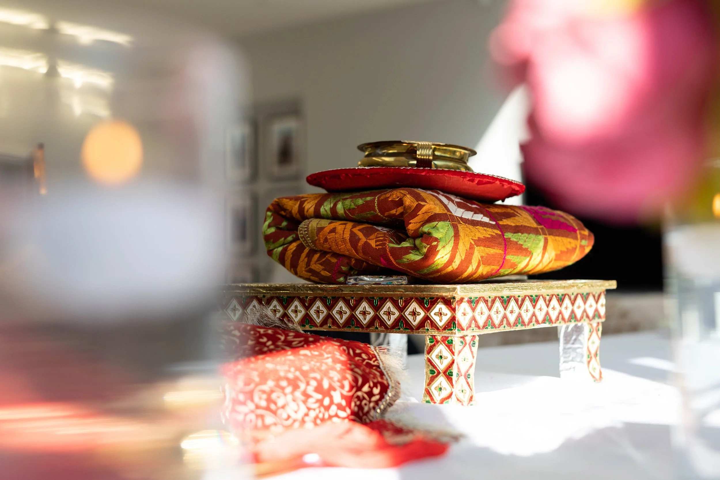 A decorated table with a colorful fabric wrapped object, a red cushion, and a small ornate table with a patterned edge