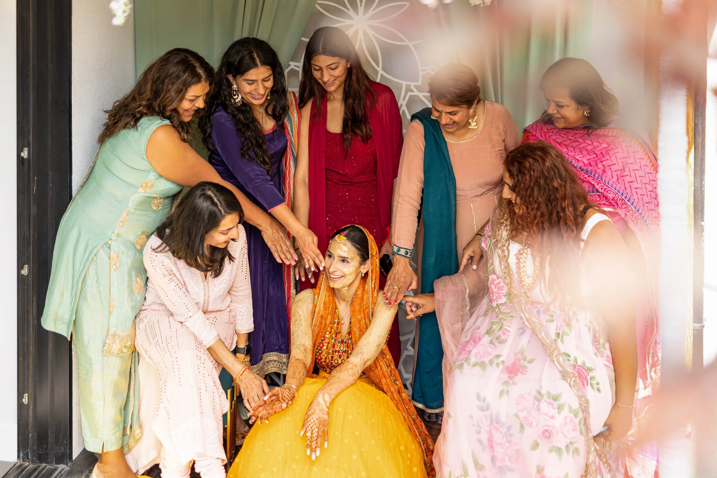 A bride in a yellow and orange traditional Indian dress is sitting on the floor, surrounded by women celebrating her pre-wedding ceremony inside a decorated indoor space.
