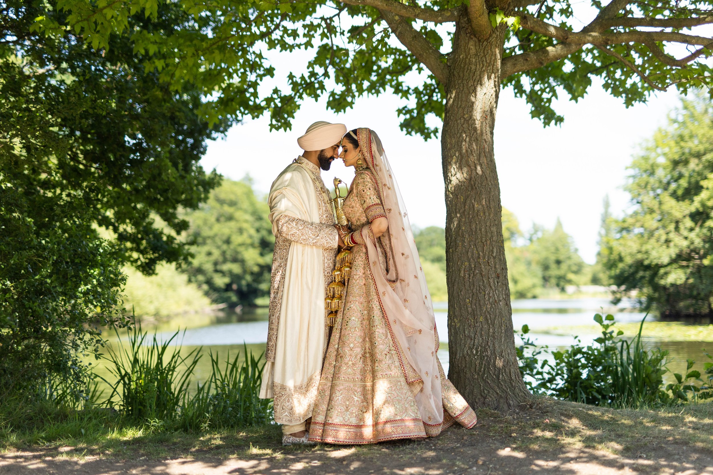 An Indian bride and groom in traditional wedding attire standing close under a tree near a lake, touching foreheads in a romantic moment.