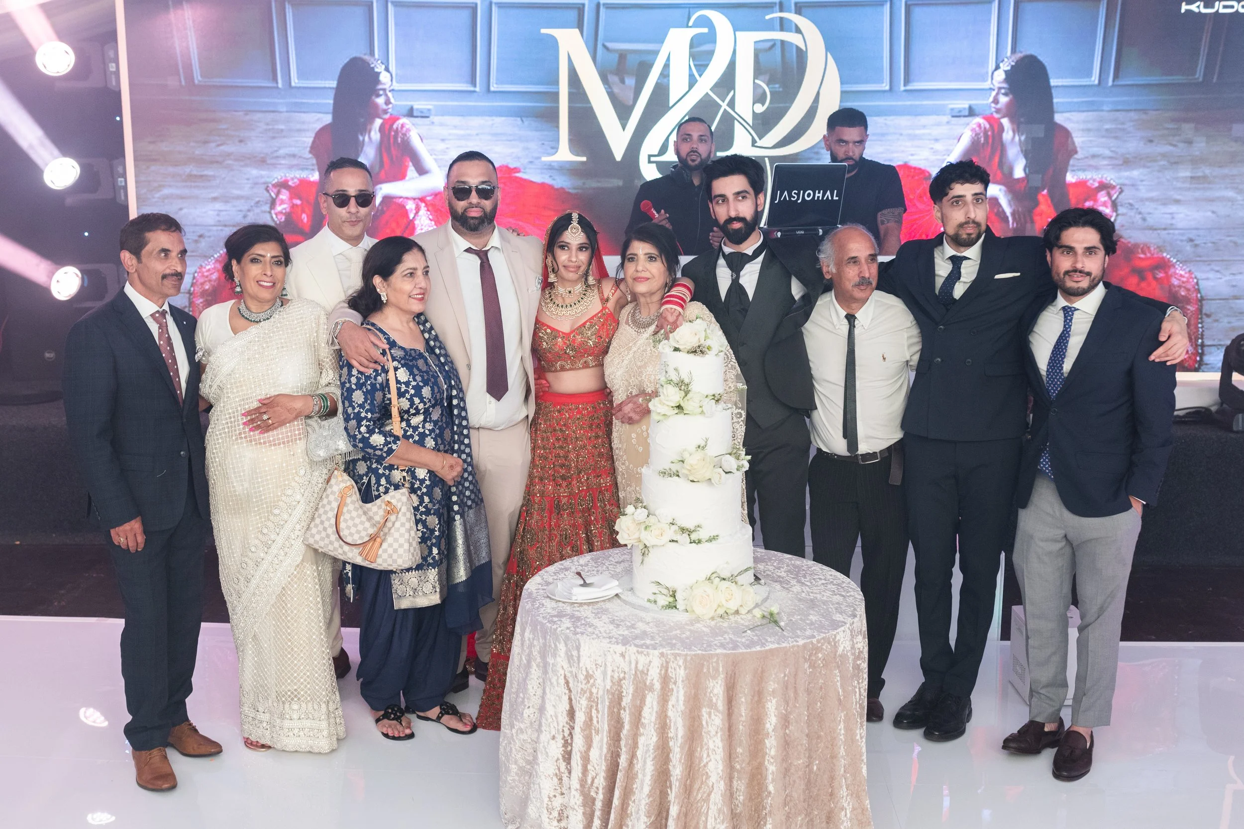 A group of people celebrating a wedding, standing around a wedding cake, with a large digital screen displaying a woman in red behind them.