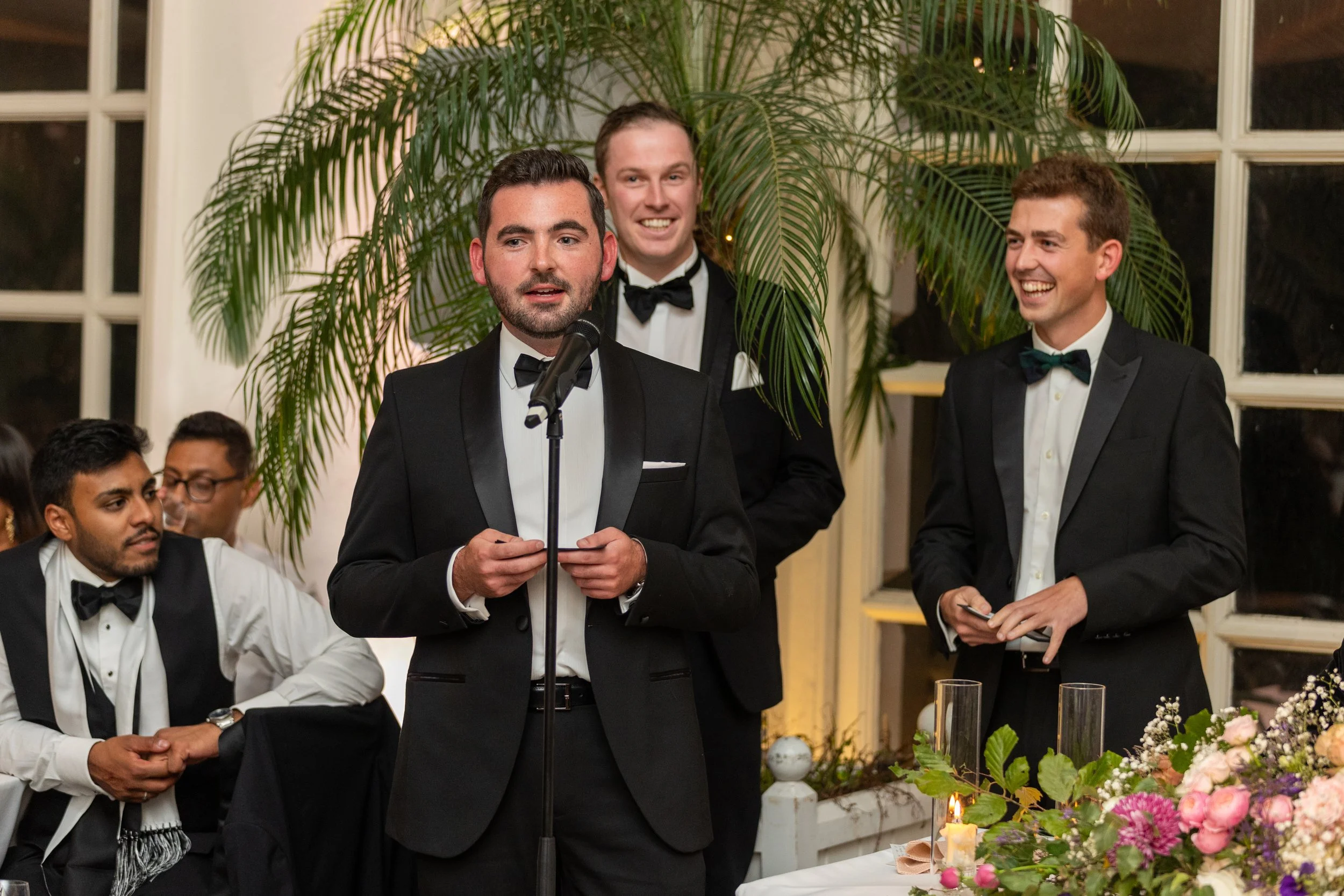 A man in a tuxedo giving a speech at a formal event, surrounded by other men in tuxedos, with a large green plant and windows in the background, and a decorated table with flowers and candles in the foreground.