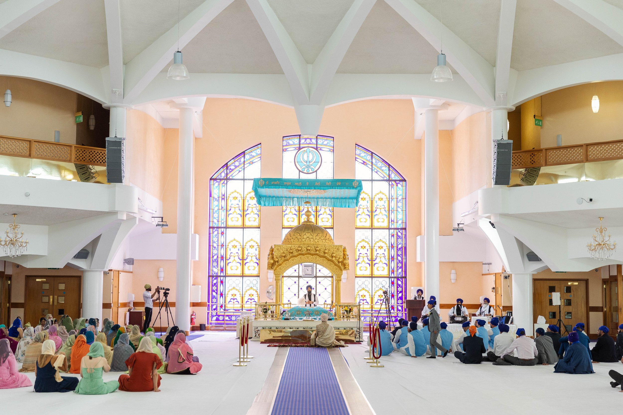 Interior of a Sikh Gurdwara with worshippers sitting on the floor, a priest at the altar, and stained glass windows in the background.