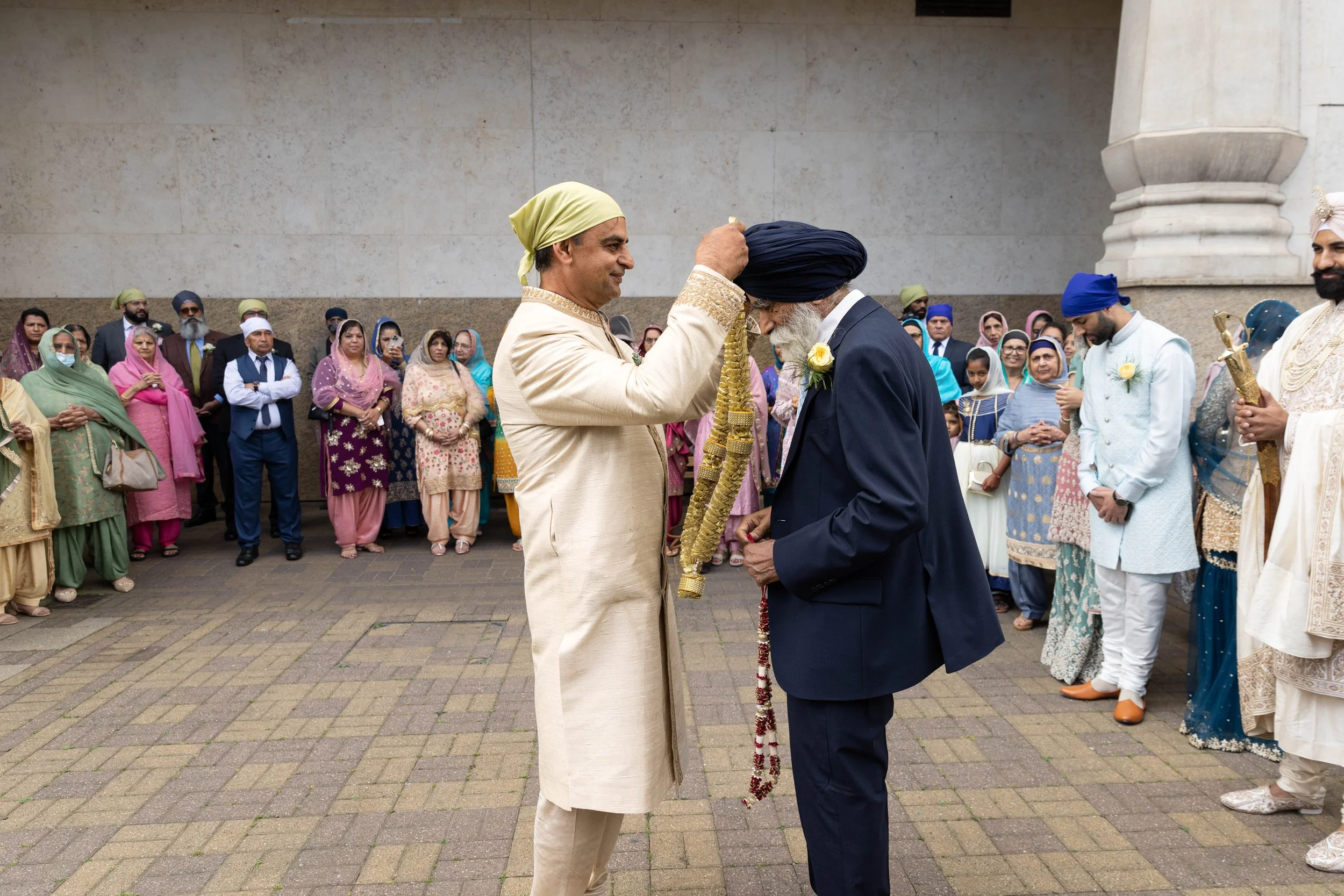 A man in traditional Indian attire placing a garland over an elderly man wearing a turban during a wedding ceremony, with guests in colorful outfits watching.