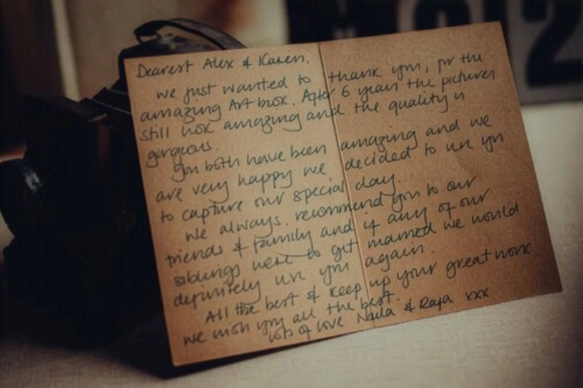 Handwritten thank you note on brown paper, placed beside a camera. The note expresses happiness and gratitude to Alex and Karen for the gift of a camera and photographs, mentioning their recommendation, and signing off with love from Nula and Rapa.