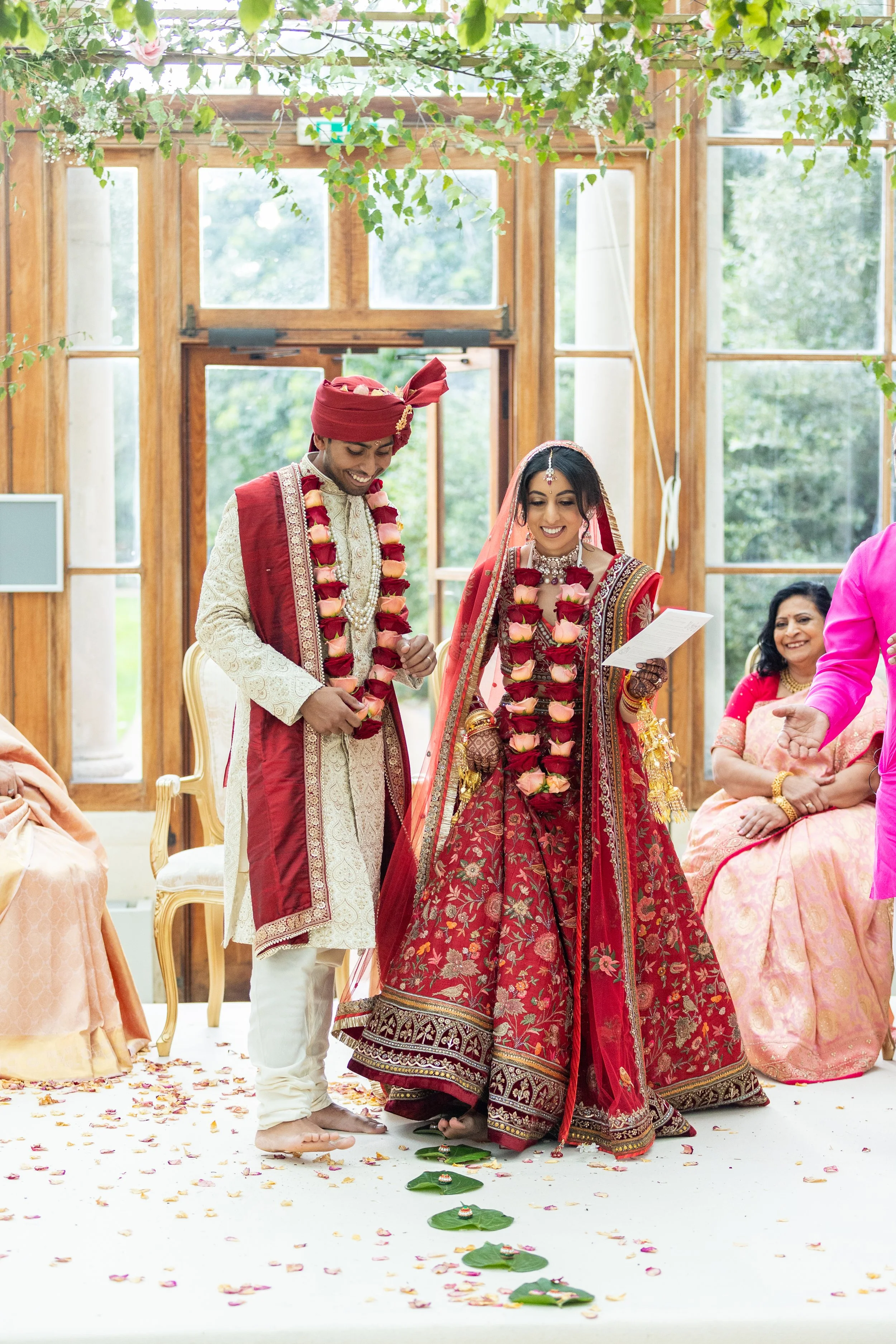 Indian bride and groom in traditional wedding attire standing on a white floor decorated with rose petals, smiling and holding hands inside a bright, wooden-walled room with large windows and greenery outside, during a wedding ceremony.