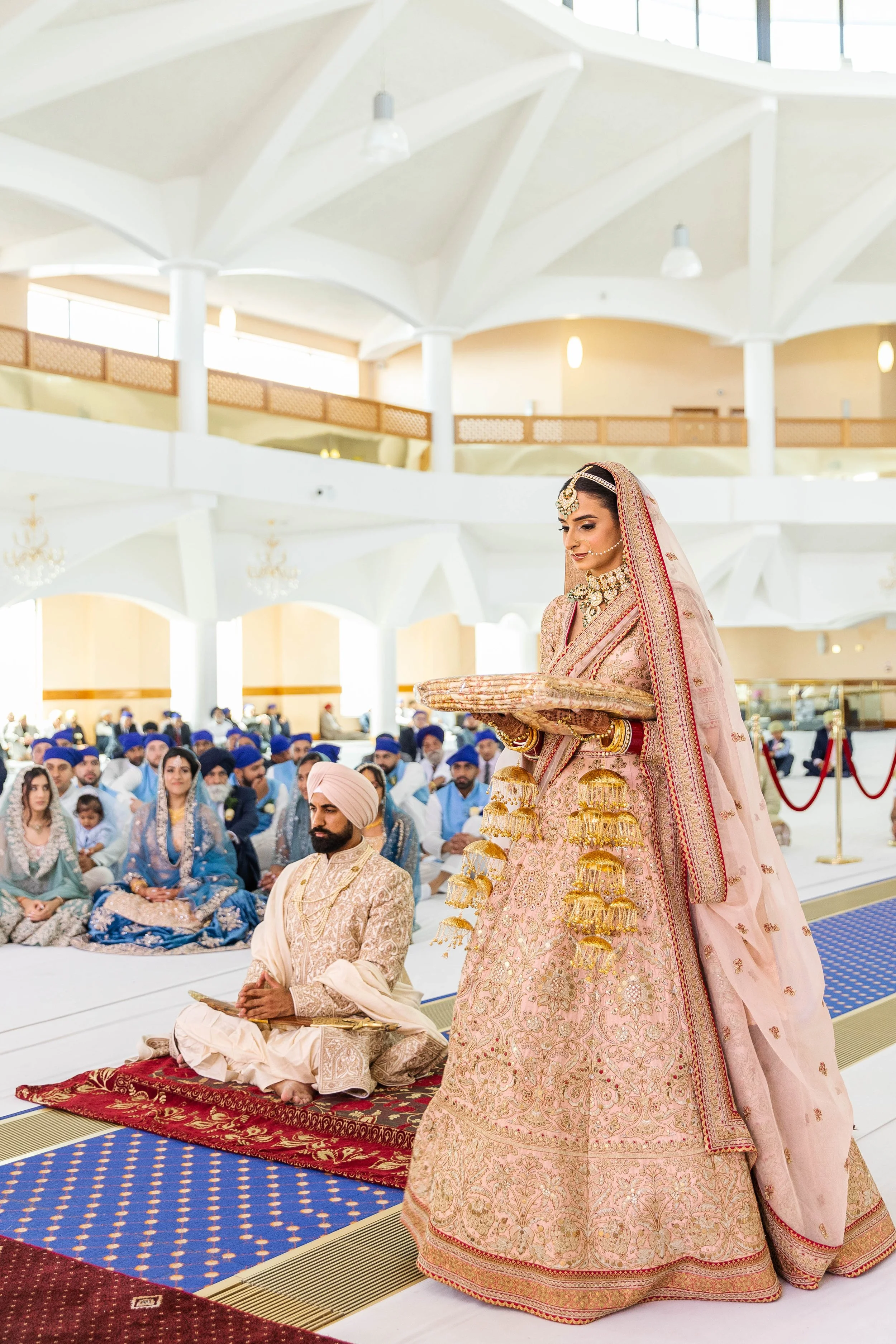Indian bride in traditional wedding attire standing with a tray, surrounded by seated guests in colorful traditional clothing, inside a spacious hall with high ceiling and chandeliers.