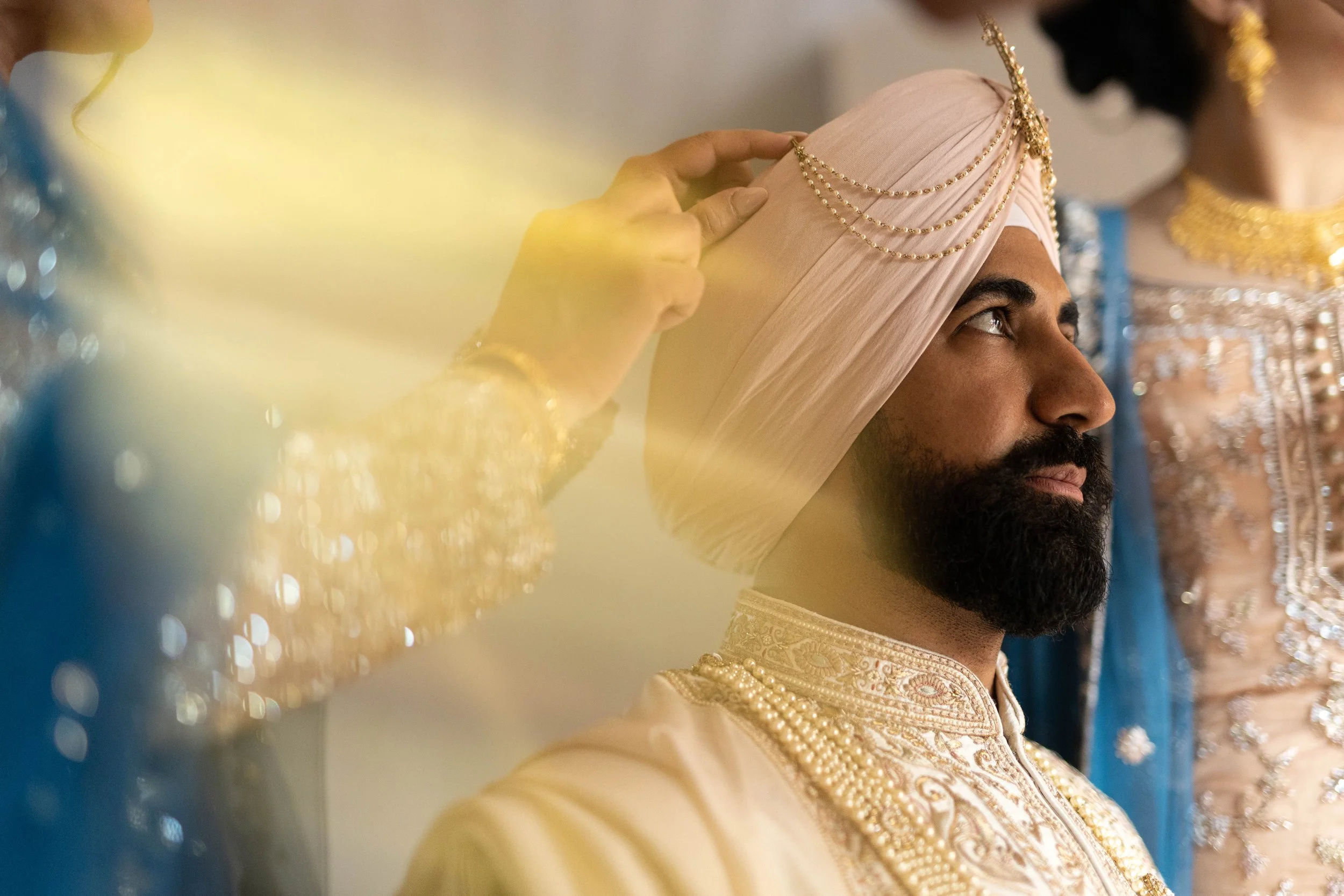 A man dressed in traditional Indian wedding attire, wearing a beige sherwani with intricate embroidery and a matching pink turban adorned with gold chains, is being assisted in adjusting his turban by a person with golden jewelry and blue clothing in