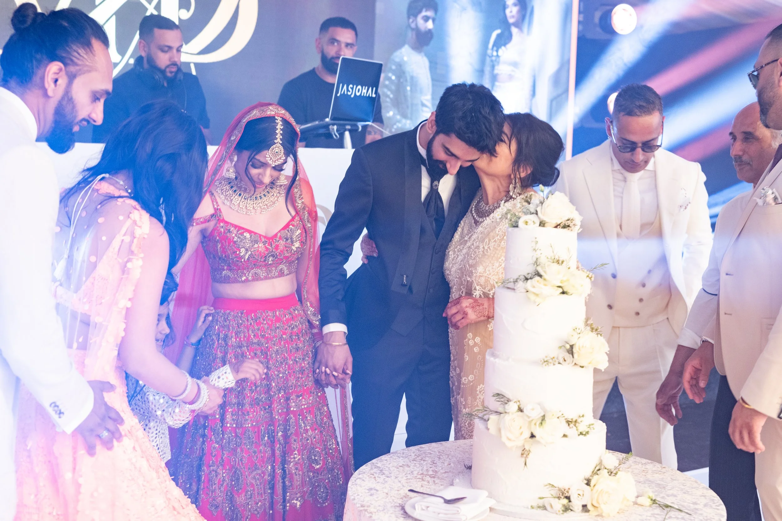 A wedding celebration with a bride and groom cutting a white tiered wedding cake decorated with white flowers. Guests in formal and traditional attire gather around, celebrating.