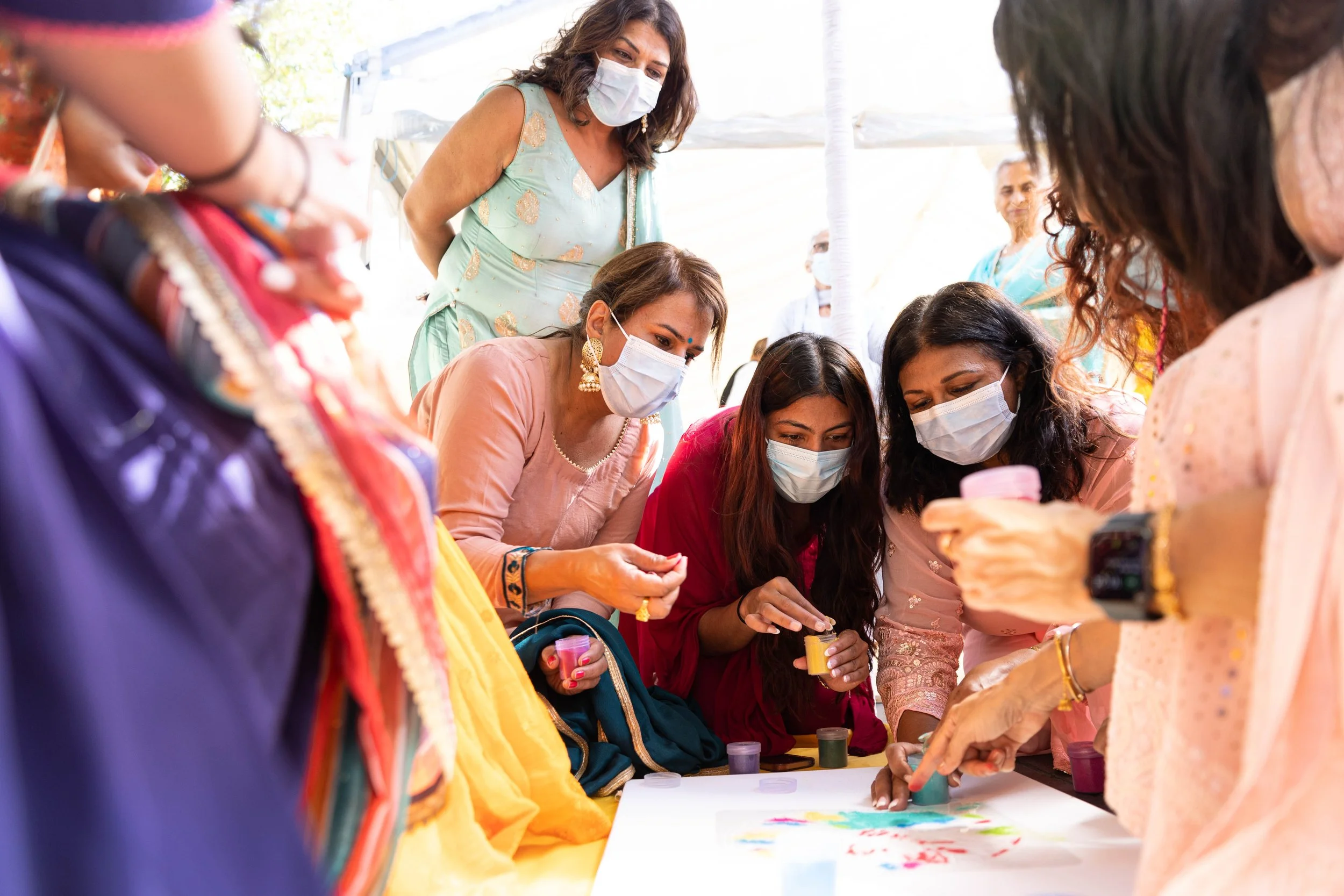 Group of women wearing masks gathered around a table, participating in a painting or craft activity. They are dressed in colorful traditional clothing, and some are holding containers of paint, focusing on their artwork.