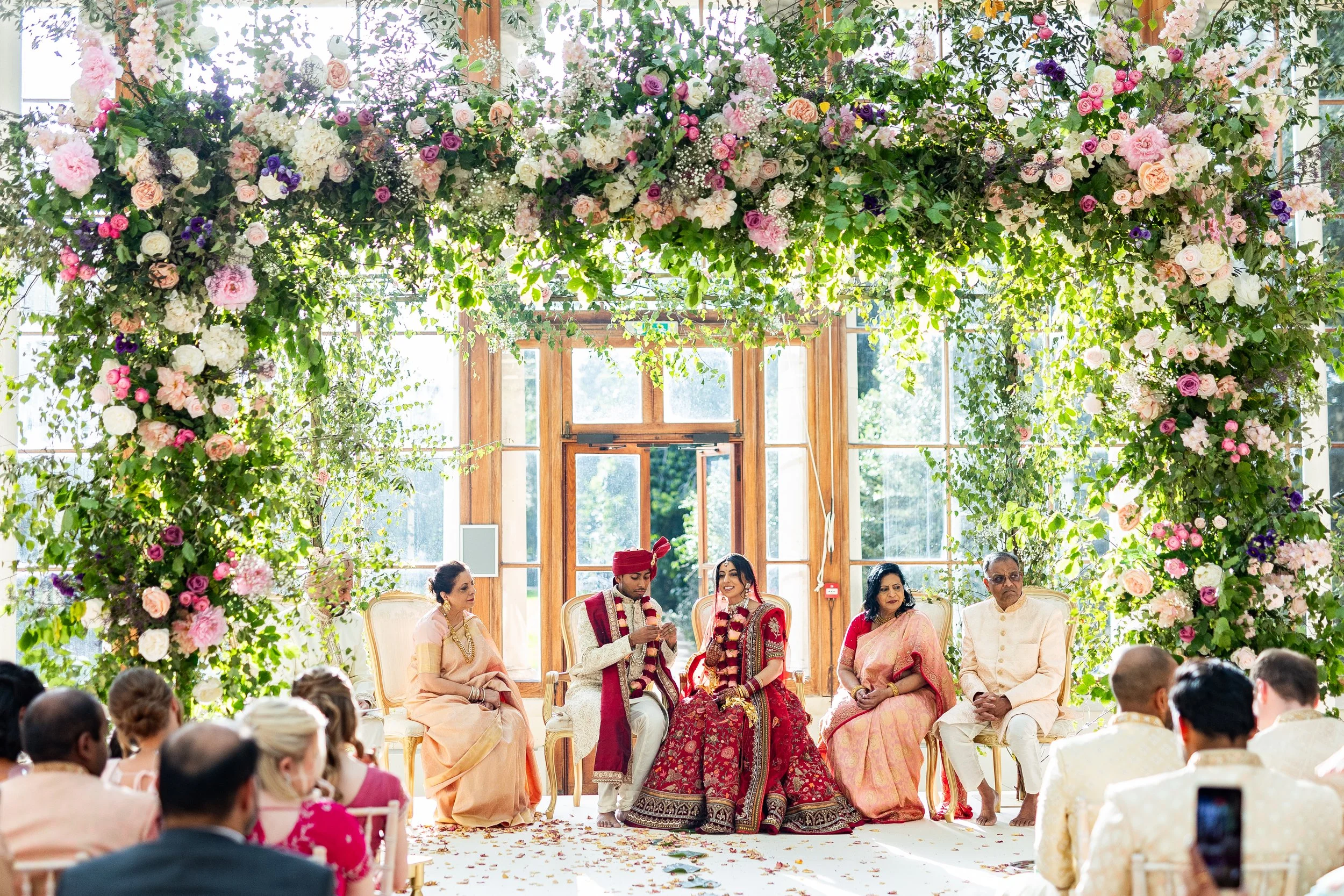 Indian wedding ceremony with a floral arch, bride and groom seated with family members, guests seated observing.