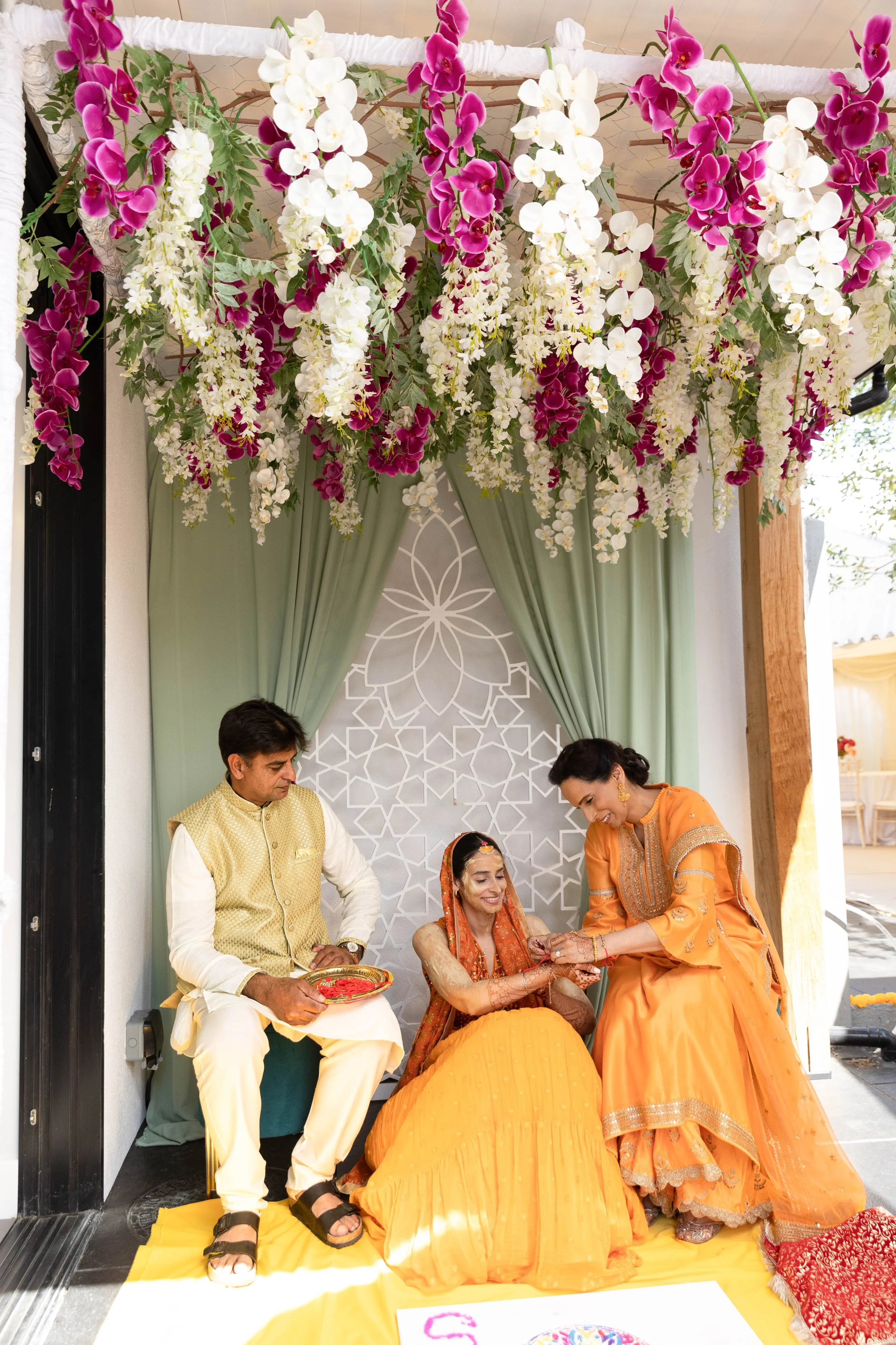 A traditional Indian wedding ceremony with a bride in a yellow dress, a groom in a cream-colored vest, and a woman in an orange sari, all seated under hanging pink and white floral decorations, with green curtains and a decorative backdrop.