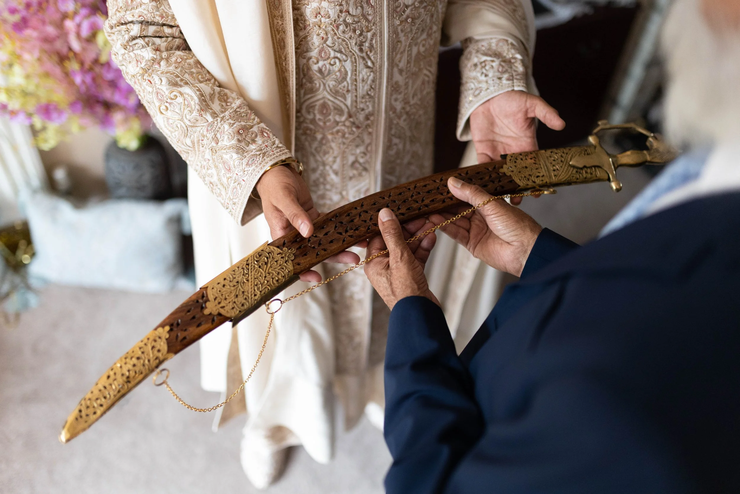 Two individuals exchange a wooden sword with gold embellishments and chain detailing during a wedding or ceremonial event. One person is dressed in traditional attire, and the other in a dark suit.