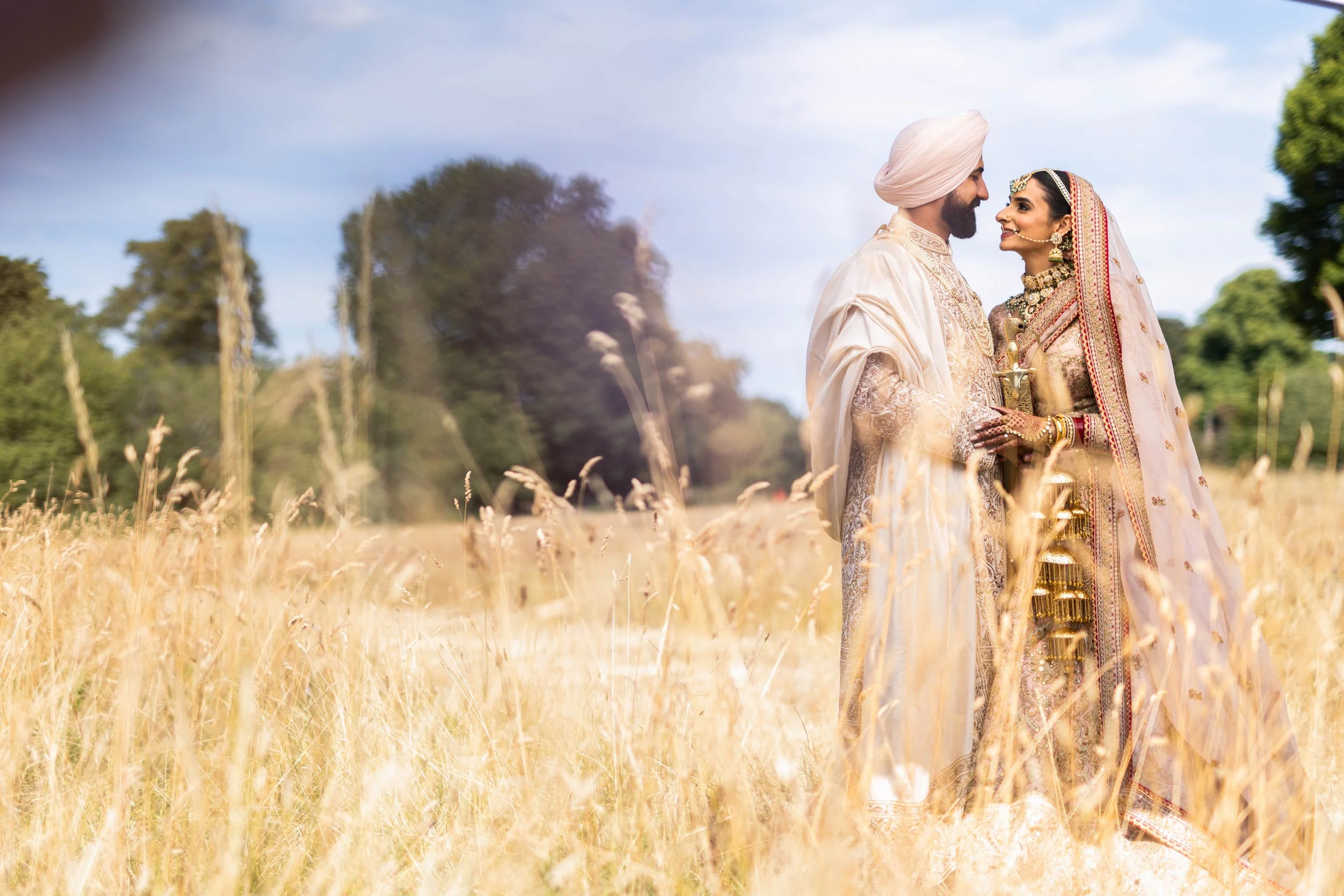 Indian bride and groom in traditional wedding attire standing in a field of tall, dry grass with trees in the background, smiling and holding hands.