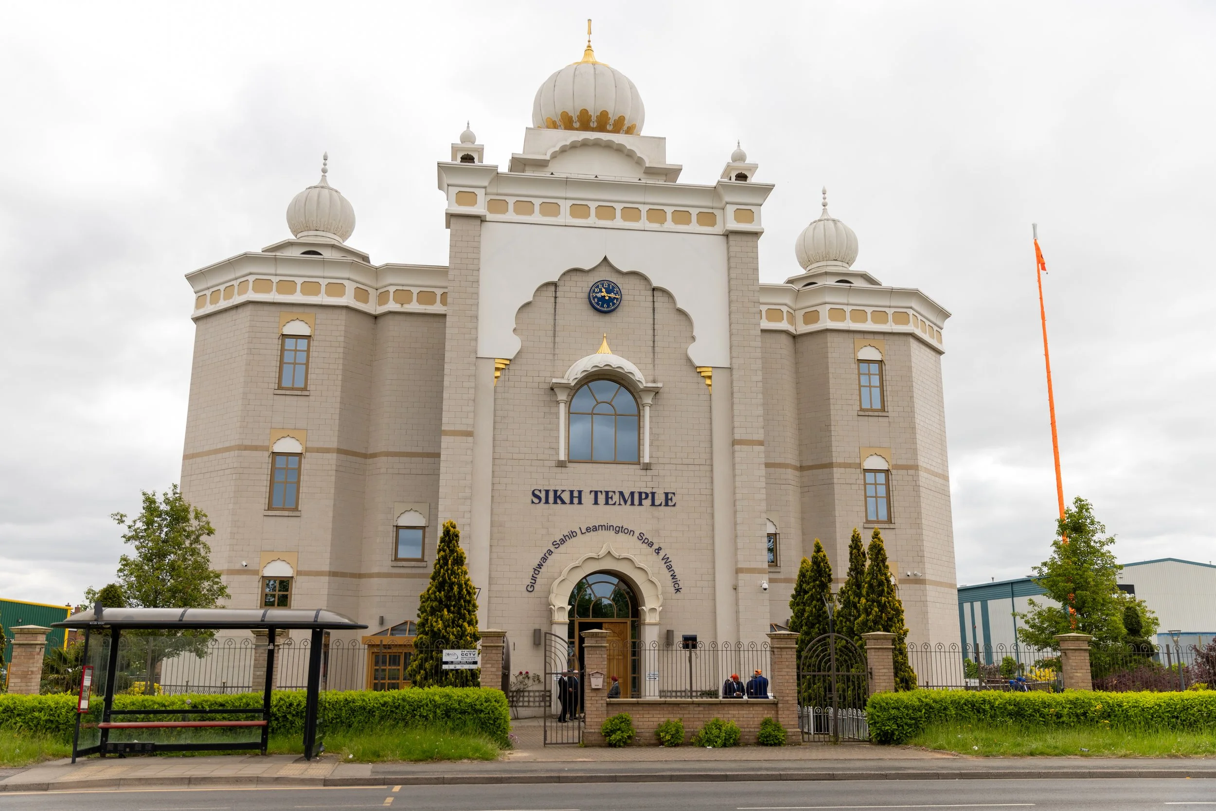Front view of a Sikh temple with beige walls, domes, and a gated entrance, surrounded by greenery and a bus stop in the foreground.