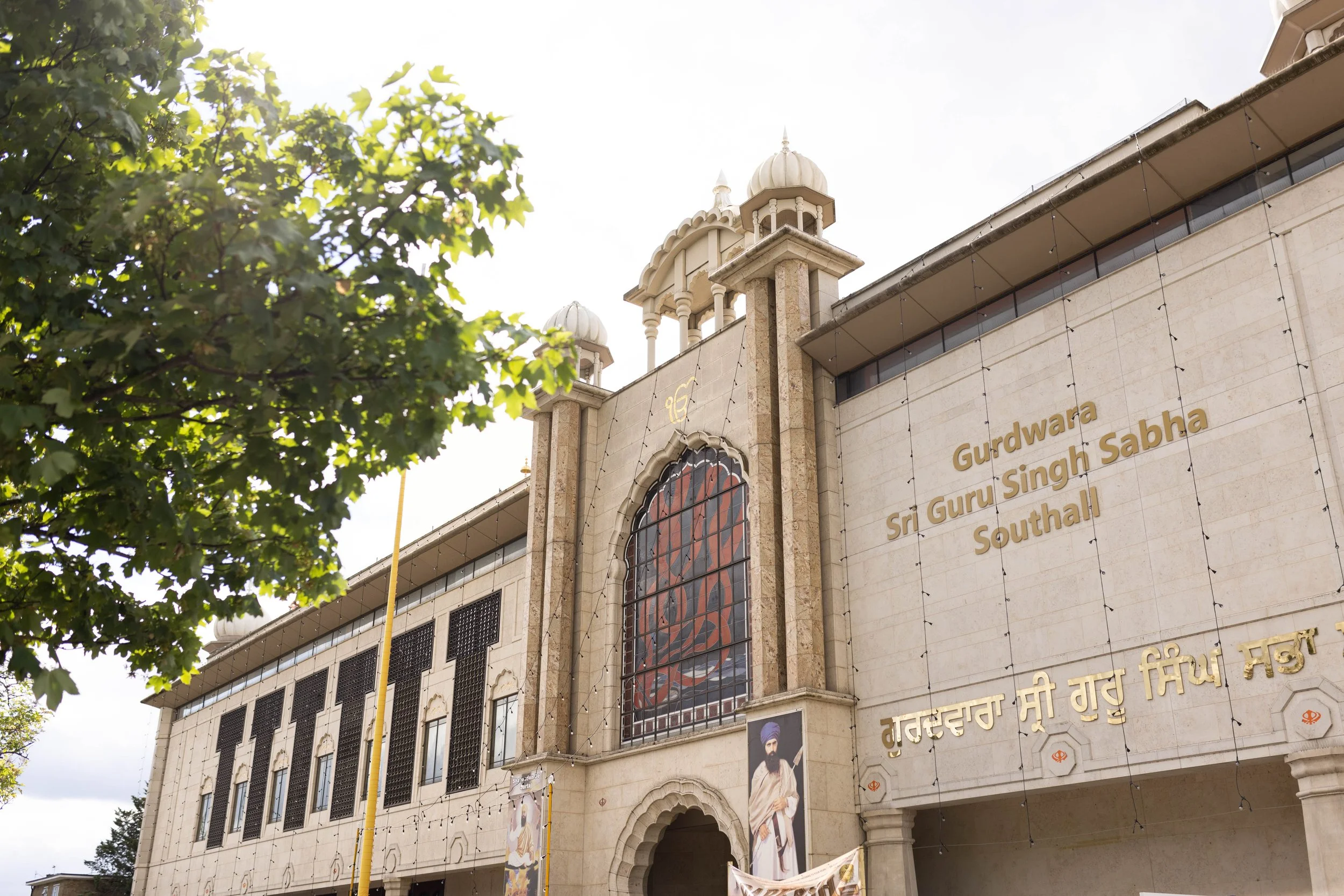 Exterior of Sri Gurdwara Guru Singh Sabha Southall with a large window, a portrait of a Sikh man in traditional attire, and gold lettering on the building's facade.