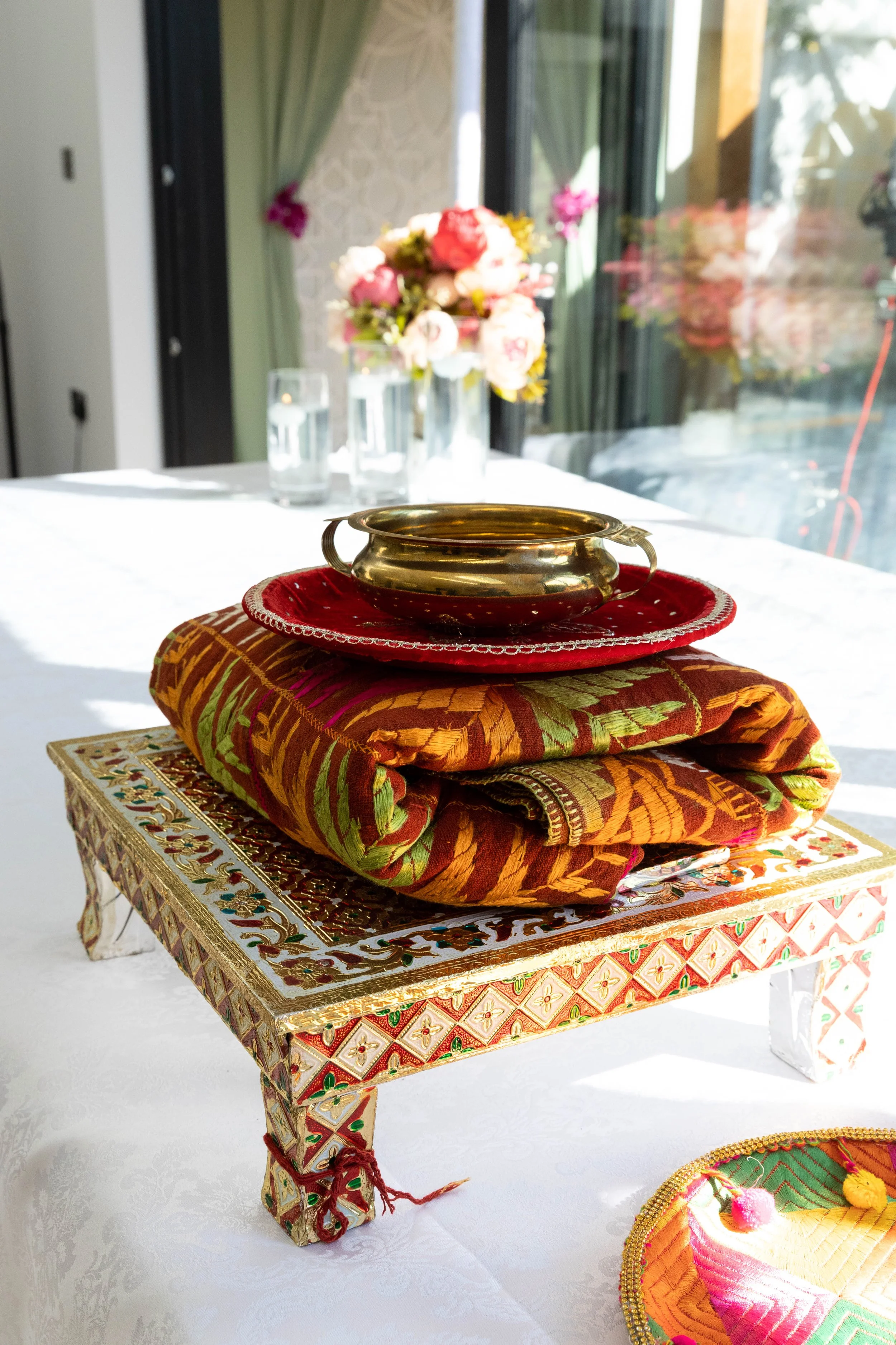 A decorative altar with a gold bowl on top, placed on a folded, colorful embroidered fabric, all on an ornate, painted wooden stand. In the background, there is a vase of pink flowers, a glass of water, and a window with curtains.