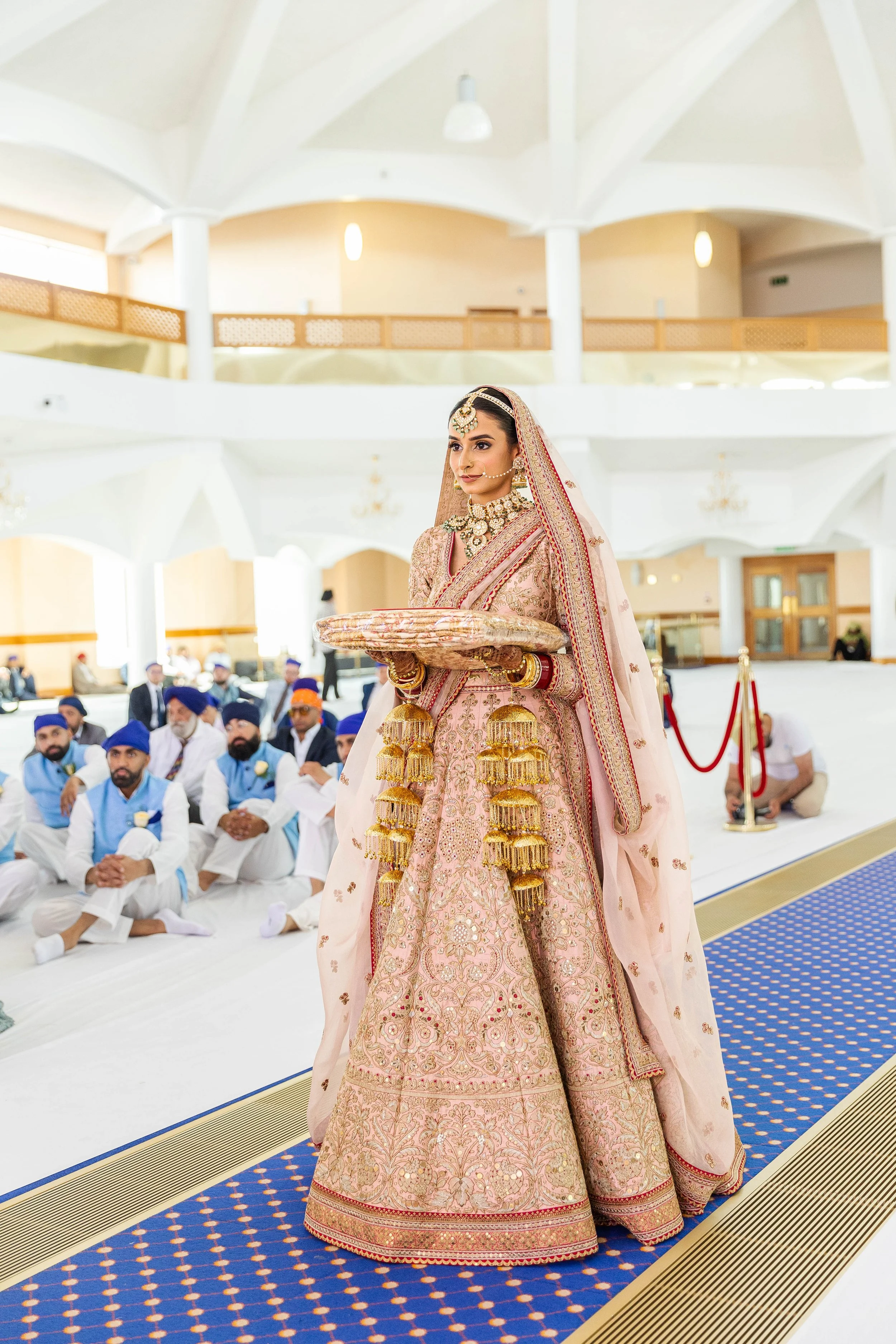 Indian bride in traditional attire holding a tray during a wedding ceremony, surrounded by seated guests.
