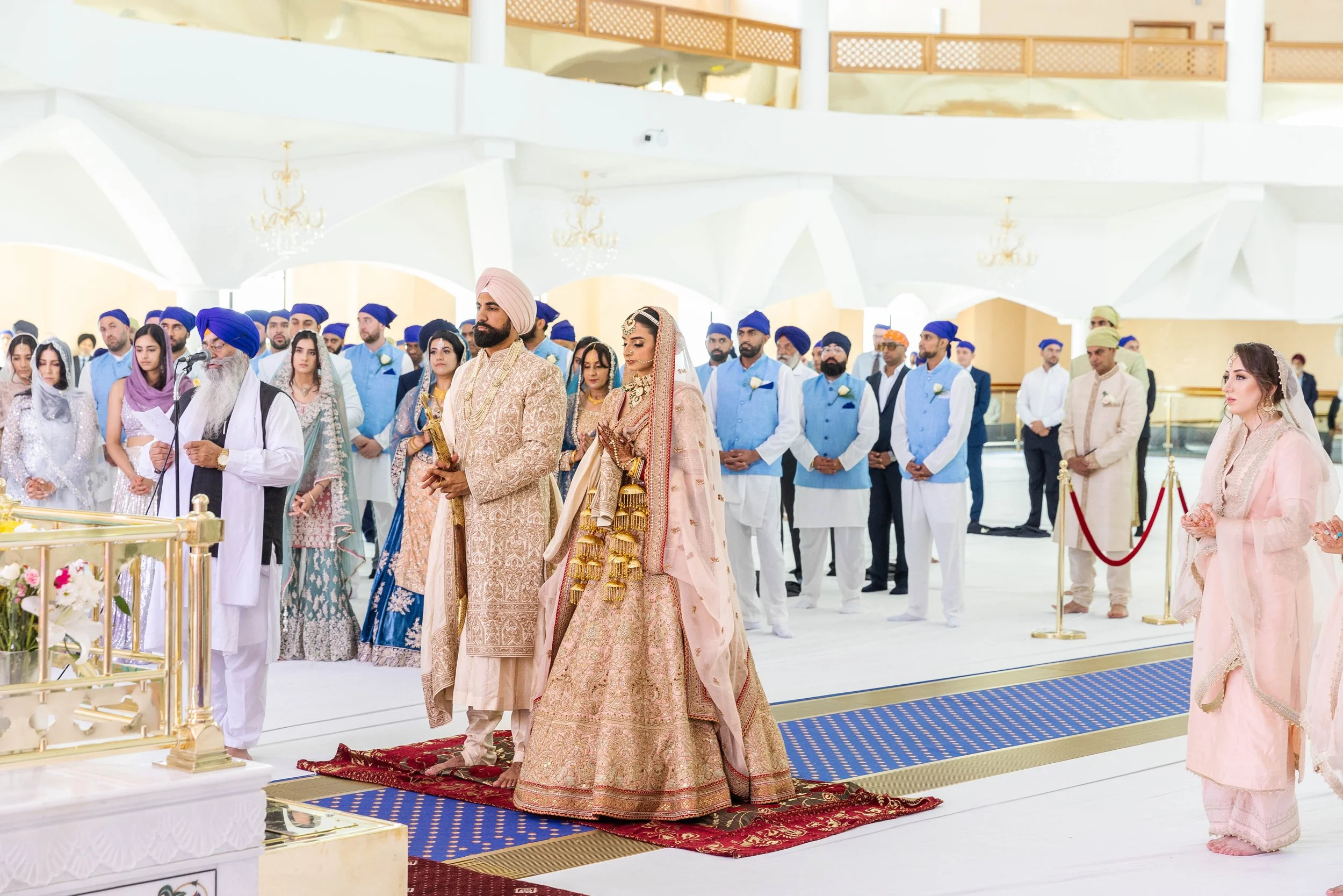 A traditional Sikh wedding ceremony in a spacious, well-lit hall with guests standing and participating, including the bride and groom dressed ornate and traditional wedding attire, along with an officiant speaking into a microphone.